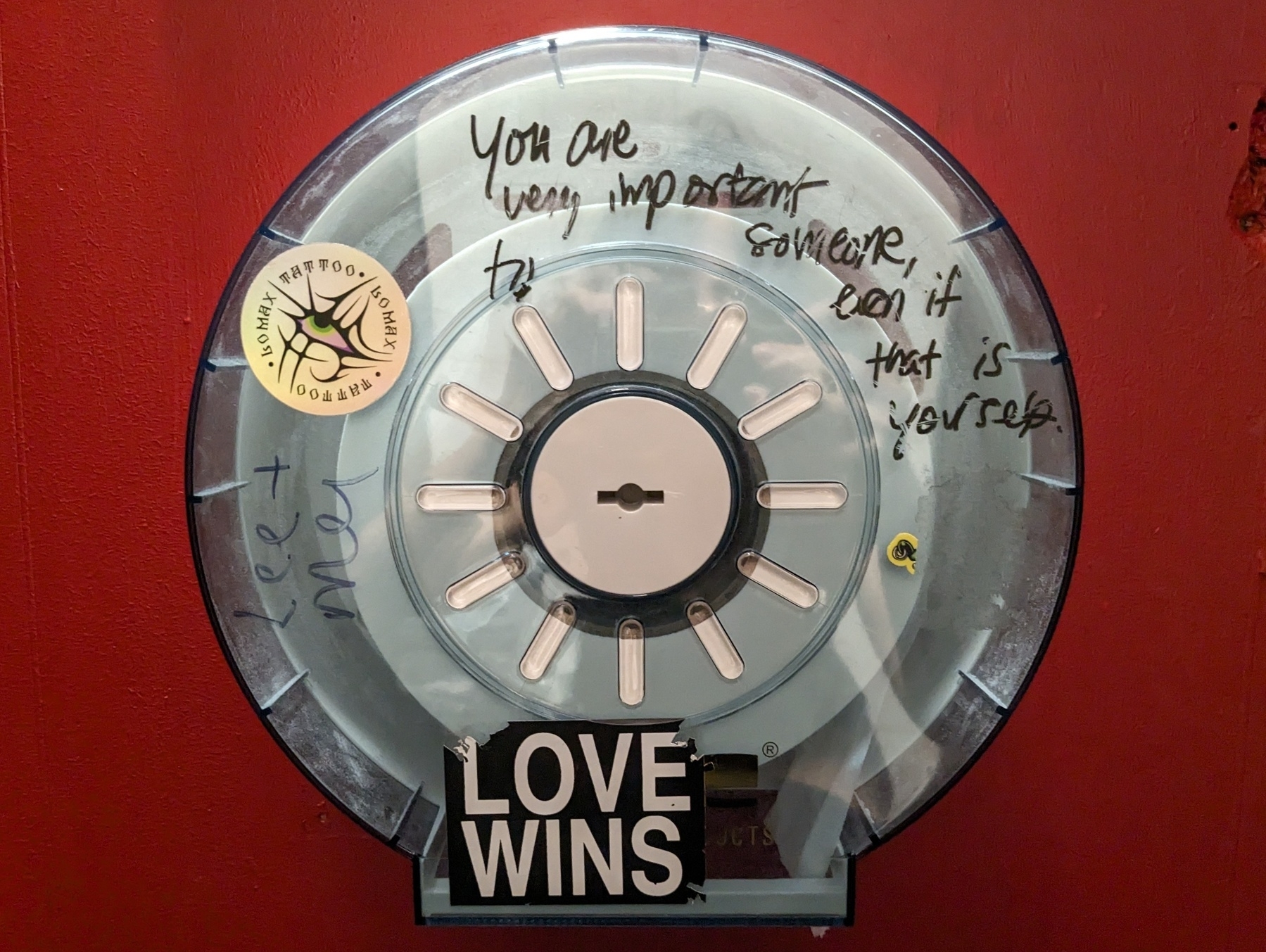 Photo of a toilet paper holder with graffiti on the back of a red door that says "you are very important to someone, even if that is yourself" and "love wins"