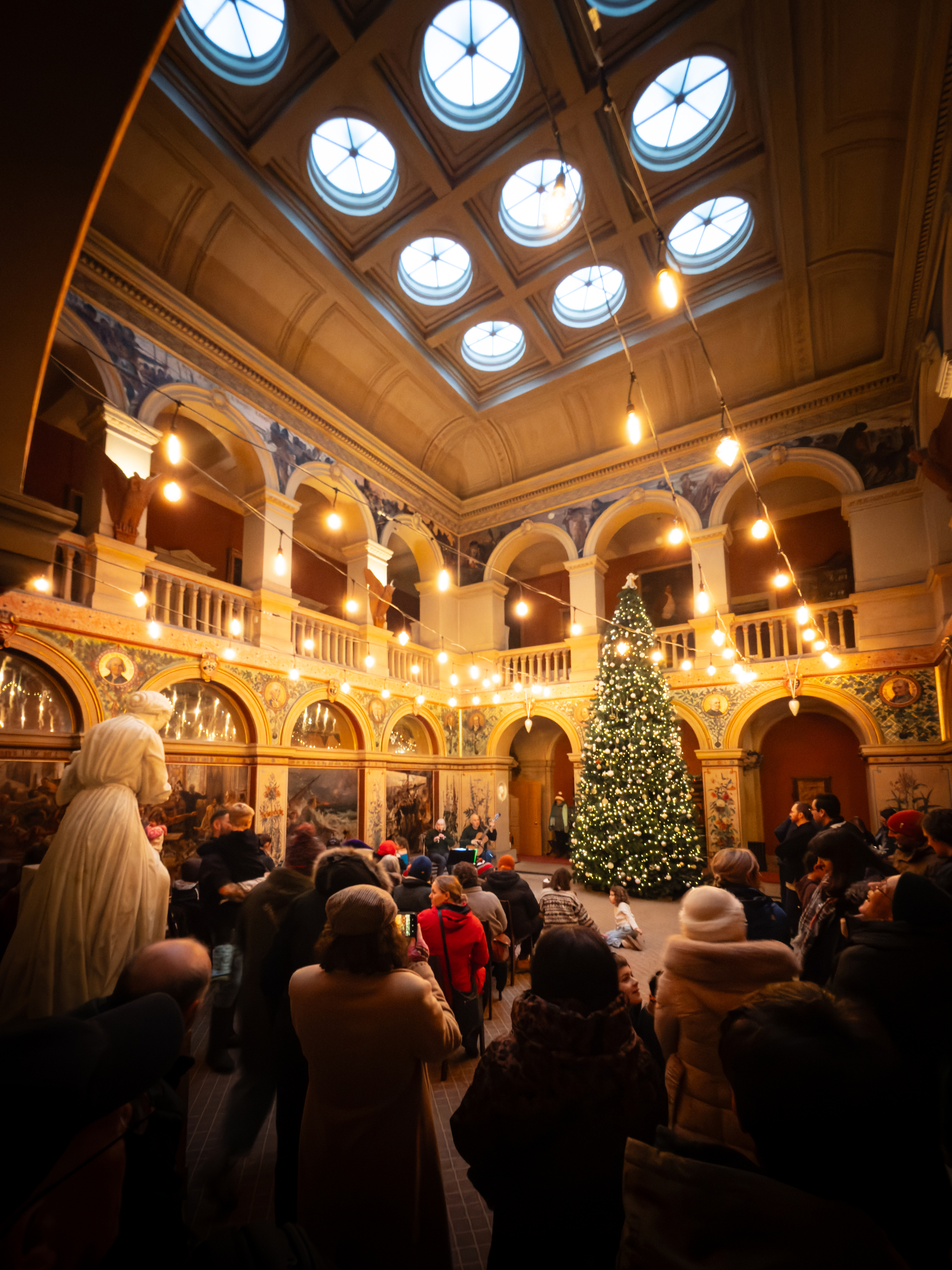 People gather in a large hall with a Christmas tree listening to musical performers at the front 