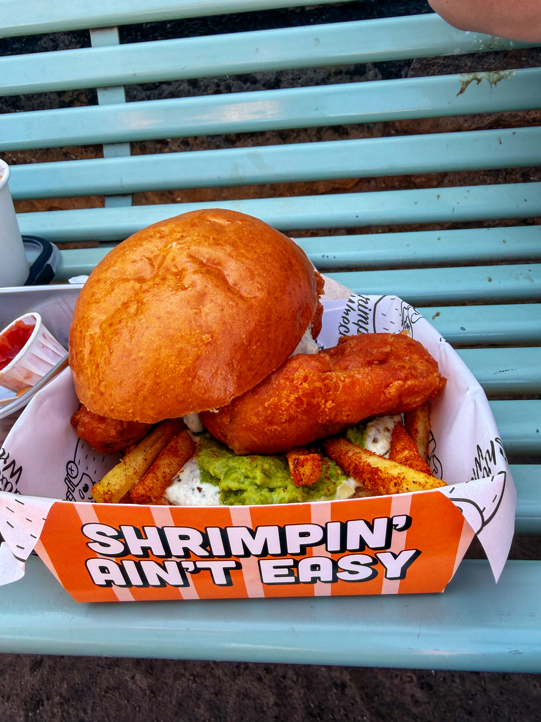 A sandwich featuring a large piece of fried fish, fries, and a spread of mushy peas is served in a branded orange box labeled Shrimpin' Ain't Easy.