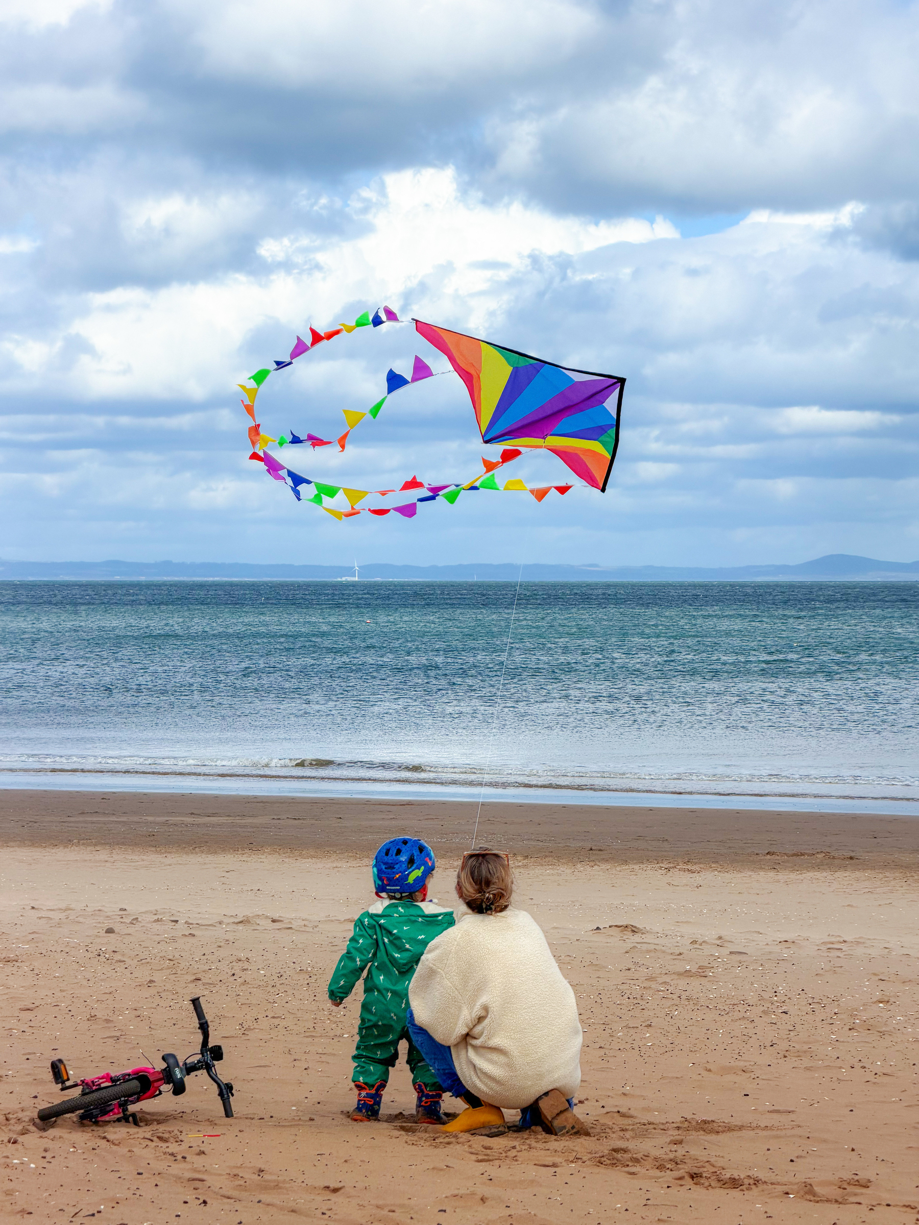 A child and an adult are flying a colorful kite on a sandy beach, with a bicycle lying nearby and the ocean in the background.