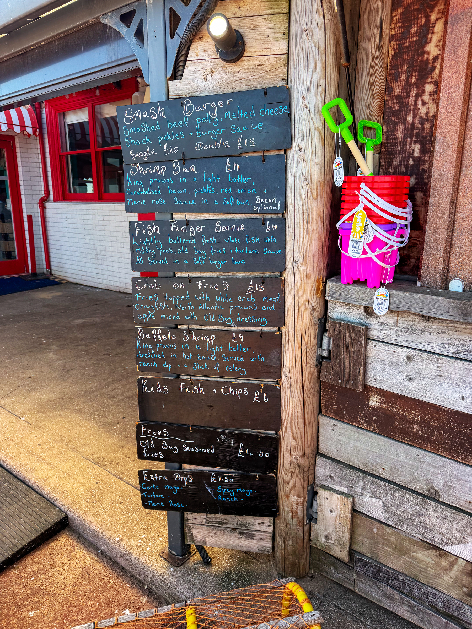 A wooden stand displays a chalkboard menu featuring seafood dishes, burgers, and fries, alongside beach toys such as buckets and shovels hanging nearby.