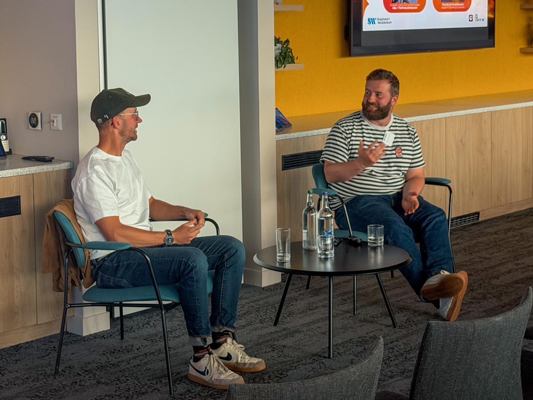 Colin and Cameron are sitting having a chat in a casual setting engaged in a conversation with a table between them holding water bottles and glasses.