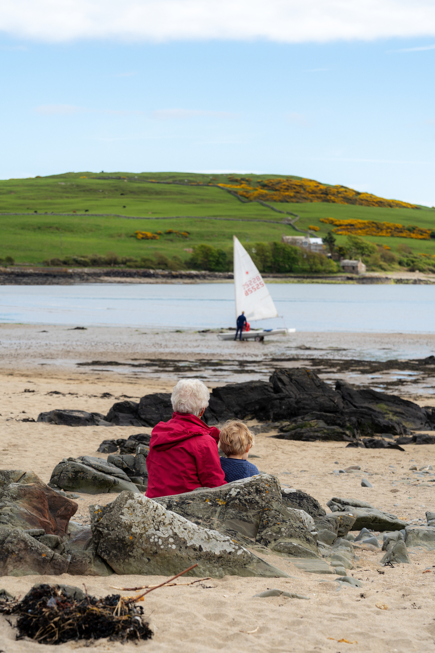 Two people are sitting on rocks at a beach, watching a small sailboat on the water, with green hills in the background.
