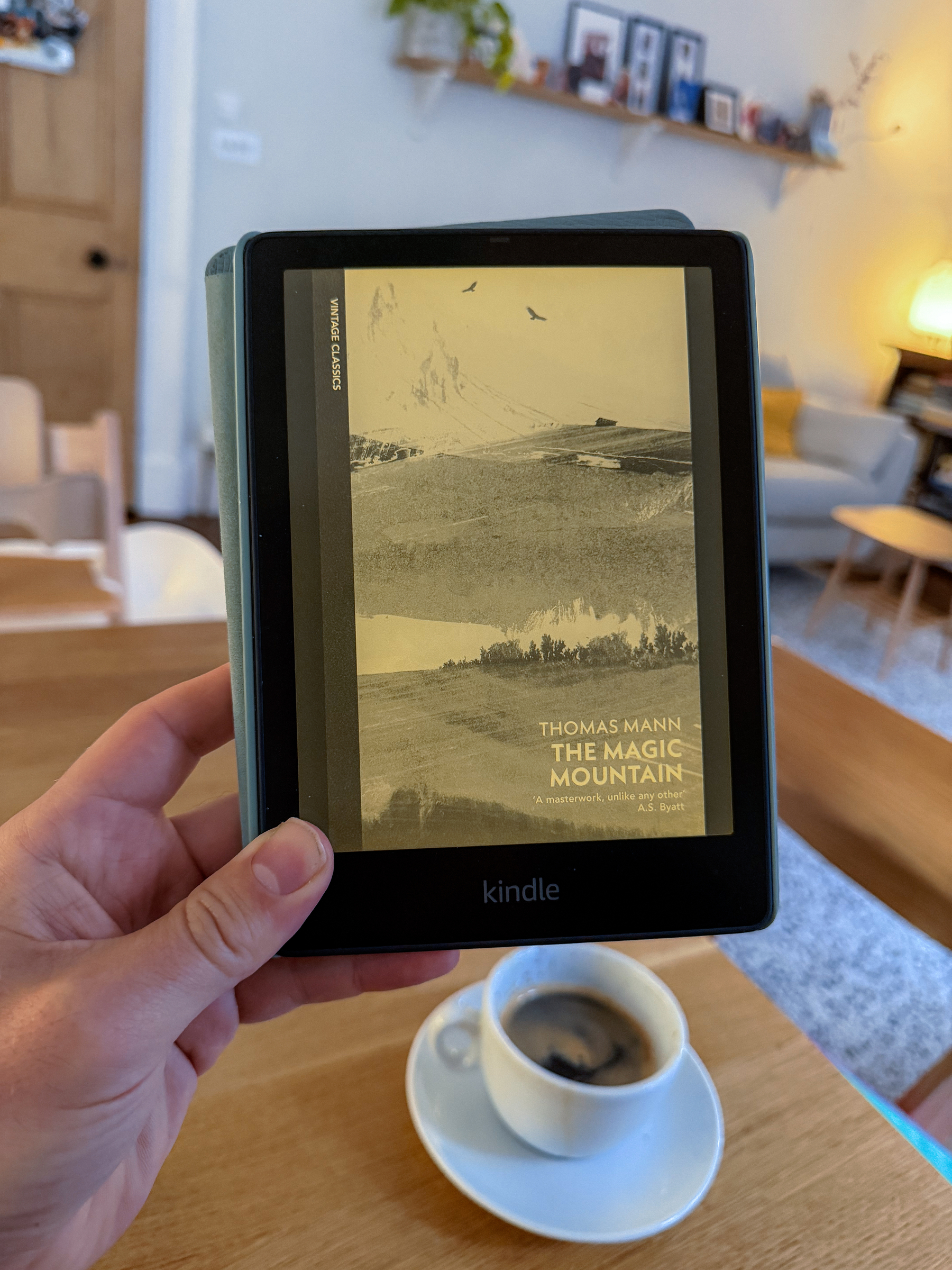  a Kindle displaying the cover of The Magic Mountain by Thomas Mann, with a cup of coffee on a table in a cozy, well-lit room in the background.