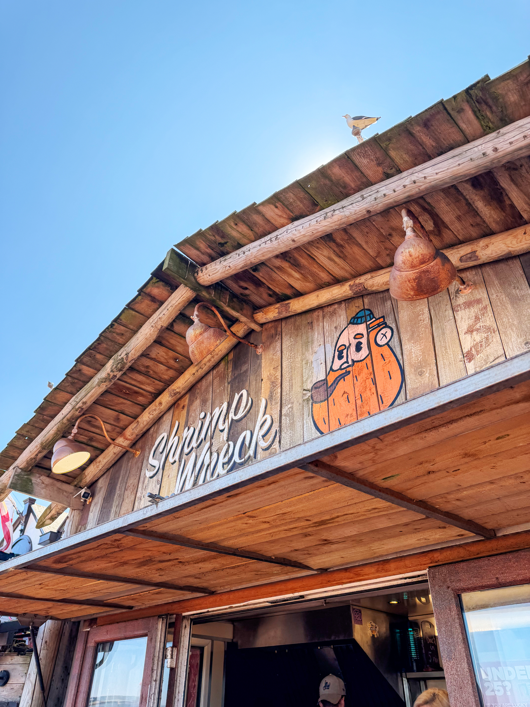 A rustic wooden seafood shack named Shrimp Wreck features two large lantern-style lights and a whimsical painted character on its front facade under a clear blue sky.