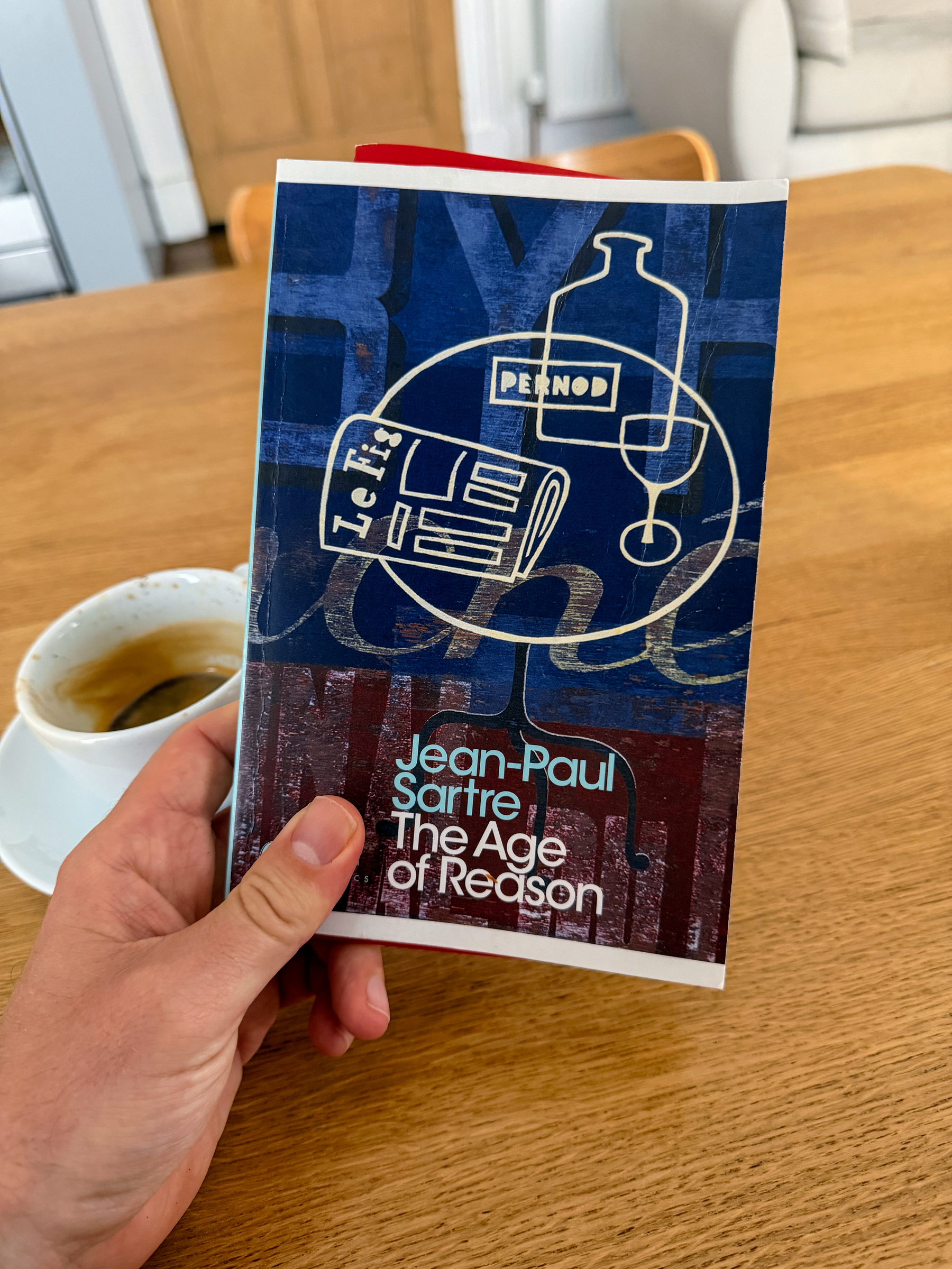 holding a copy of Jean-Paul Sartre's book The Age of Reason over a wooden table with a cup of coffee beside it.
