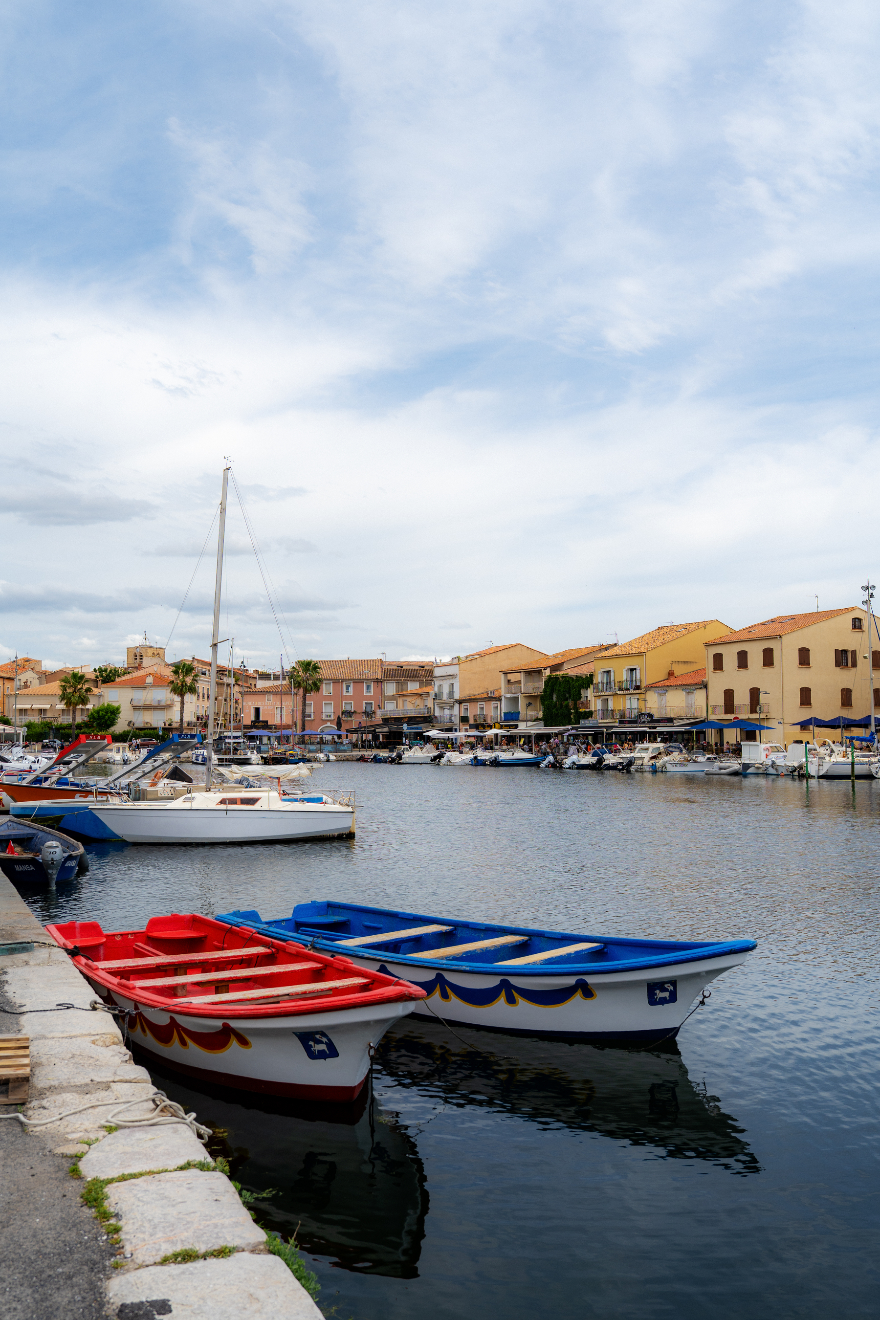 Colorful boats are docked along a waterfront with a scenic view of buildings and a cloudy sky in the background.