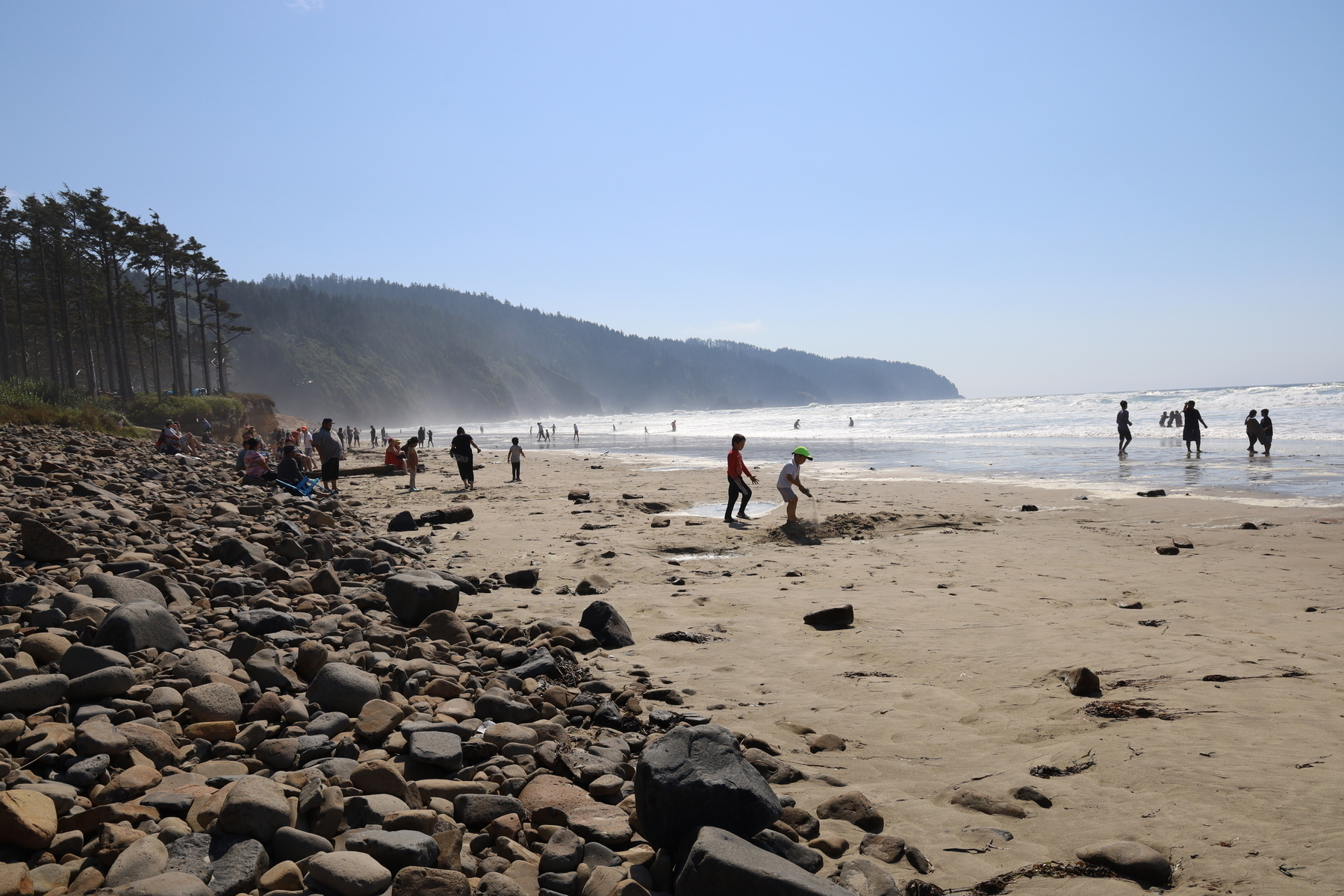 A view looking north at the Oregon coast where Cape Lookout juts out into the ocean. Childs and adults play in the narrow beach between the rocks and the ocean. 