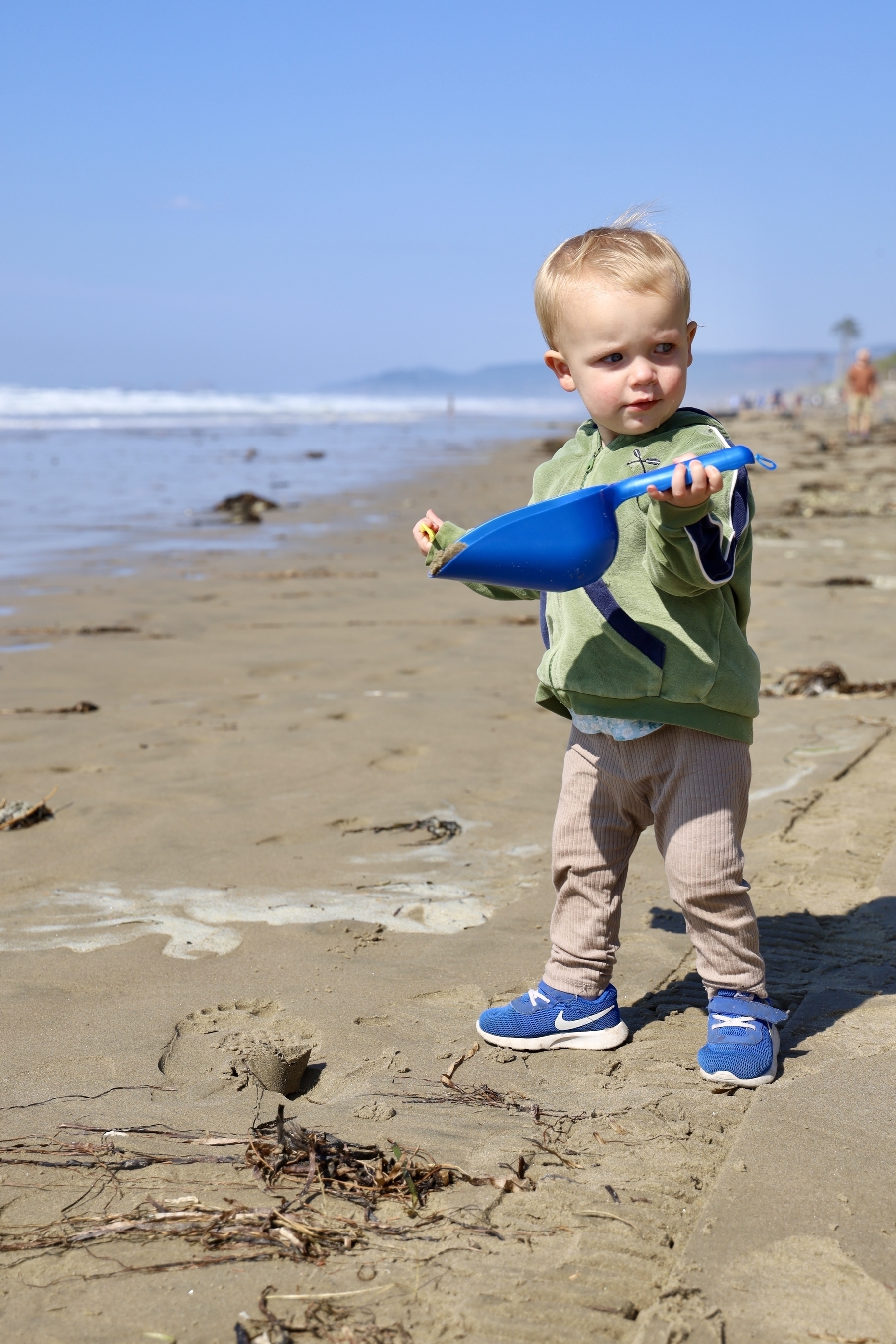A 19-month-old toddler uses a blue scooper to pick up sand at the beach. 