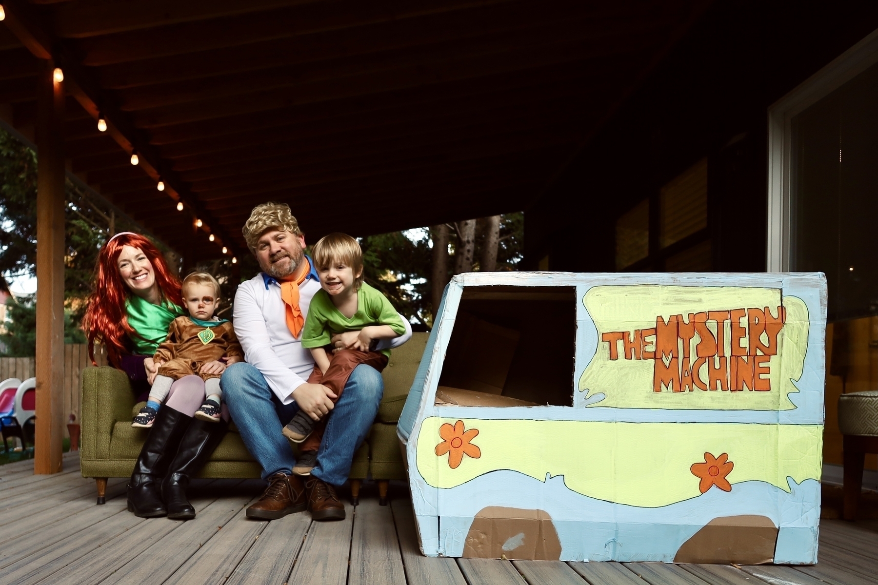 A family of four with two little kids, all dressed like Scooby doo and the gang with a cardboard box painted like the Mystery Machine. The baby is Scooby, the 4 year old is Shaggy, the dad is Fred and the Mom is Daphne. 