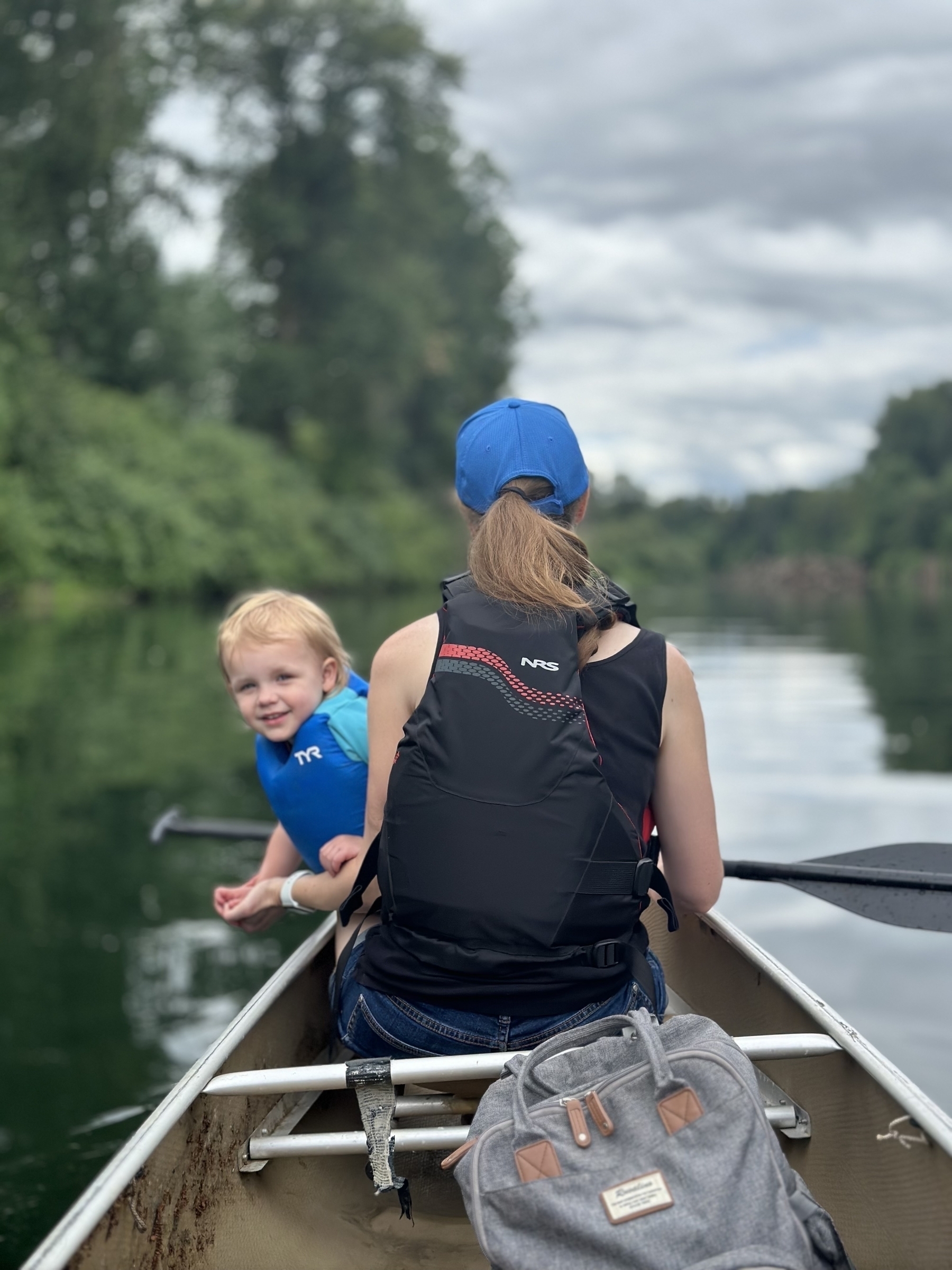 A person wearing a blue cap and a black life jacket is sitting in a canoe paddling on a calm river, with a smiling child also in the canoe surrounded by trees and a cloudy sky.