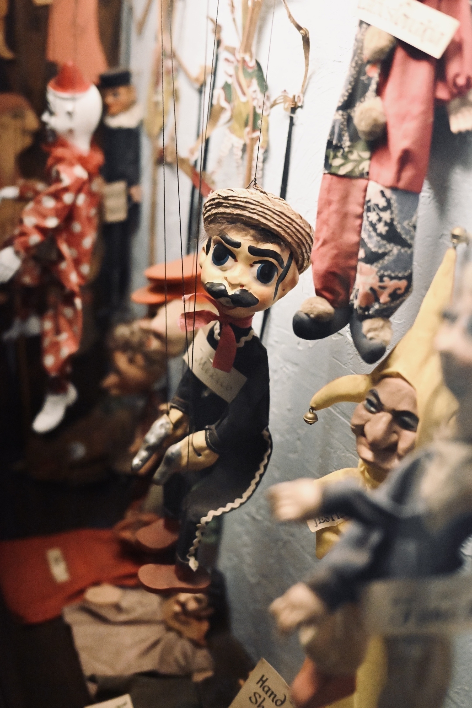 Marionette puppets with various costumes and facial expressions hang on display, some with strings attached to their limbs and labels around their necks.