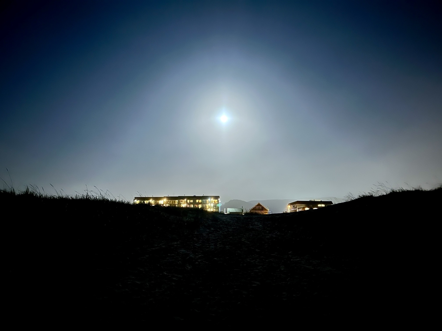 A dark coastal scene looking inland of sand dunes, and a bright, nearly full moon rising above some low-rise hotels. 