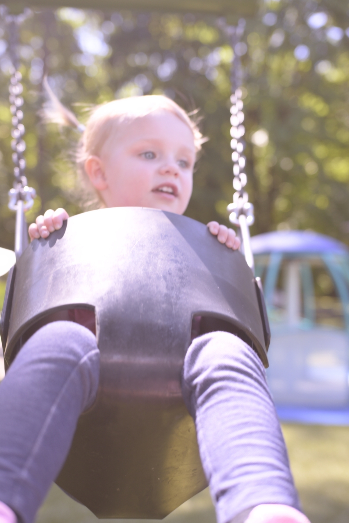 A young child with light hair is sitting in a swing, holding onto the sides, with a blurred background of greenery and playground equipment.