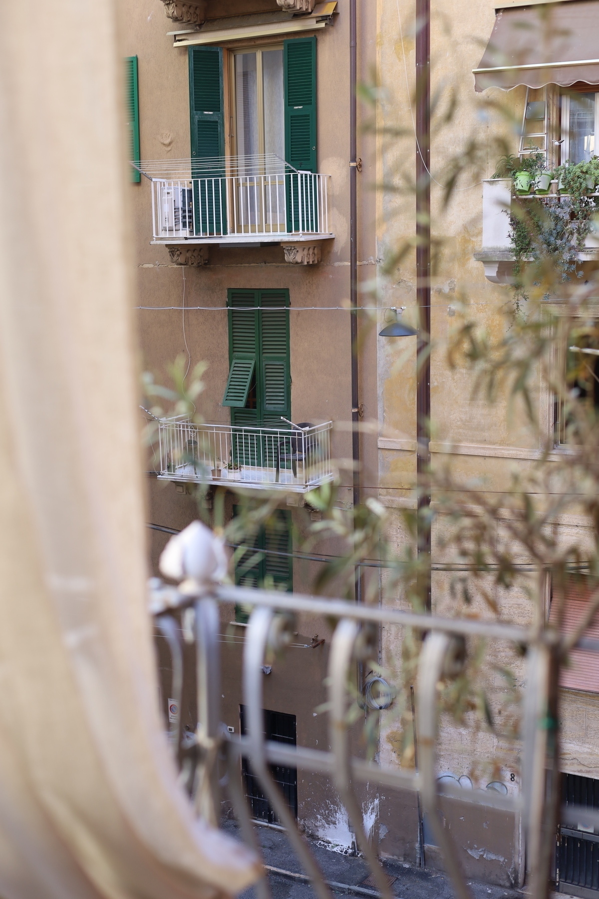 A view of a beige apartment building with green shutters and balconies is seen through a window adorned with a railing and plants.