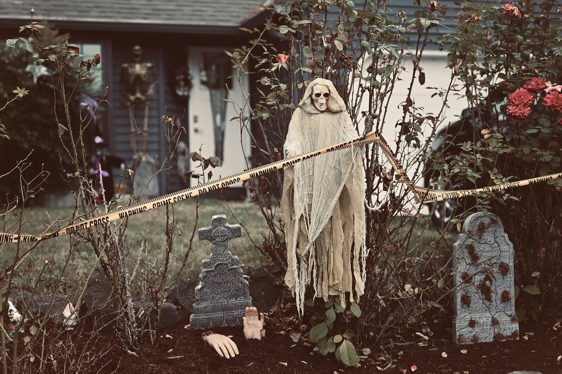 A spooky Halloween display features a ghostly figure, tombstones, and scattered body parts, surrounded by caution tape in a garden.