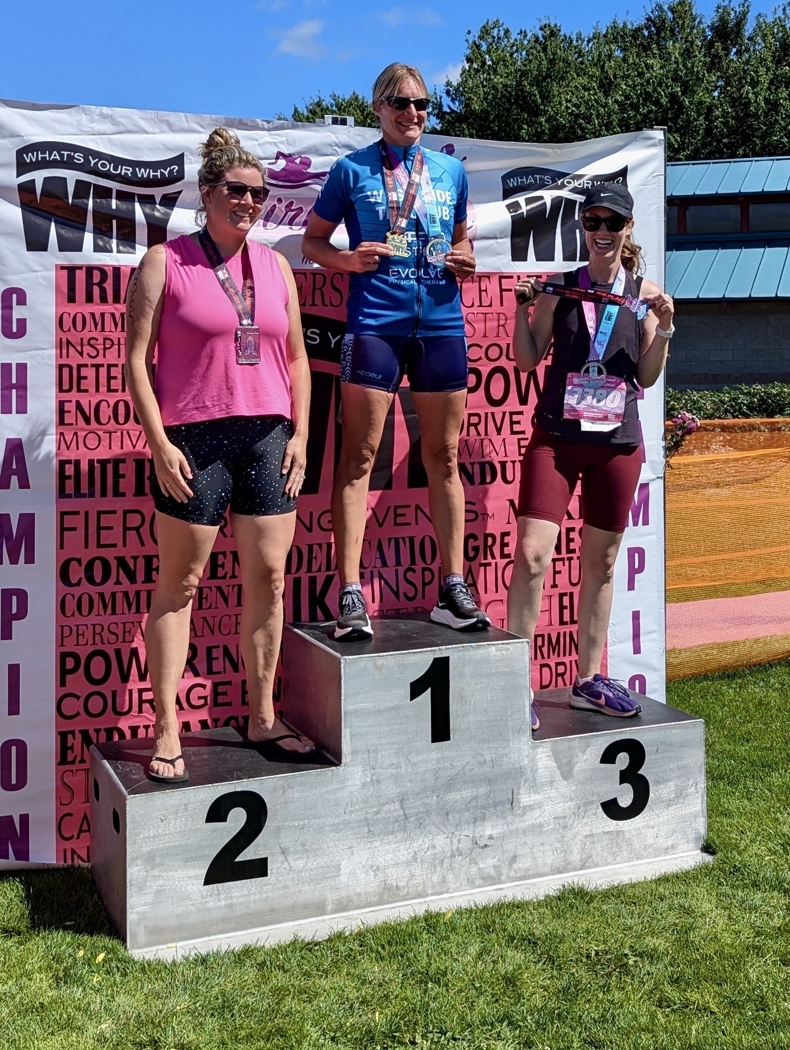 Three women wearing medals and athletic attire stand on a podium with numbered steps, celebrating a sporting event victory in front of a colorful backdrop.