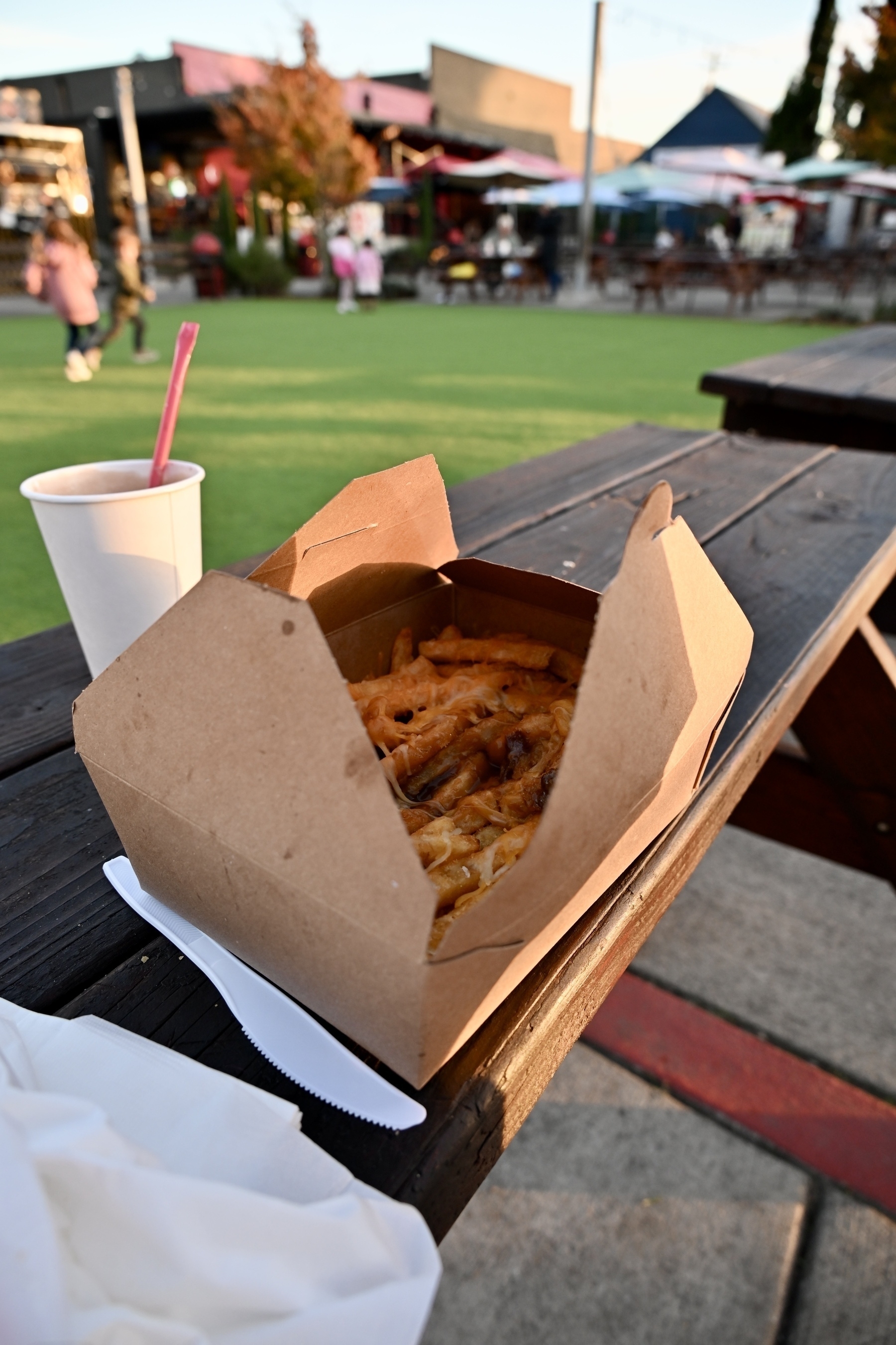 A box of fries and a cup with a straw are placed on a wooden picnic table in an outdoor setting with people and seating in the background.