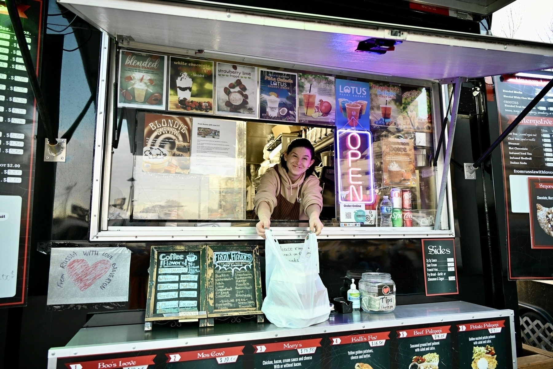 A person is serving food or drinks from a food truck displaying various menu items and an "OPEN" sign.