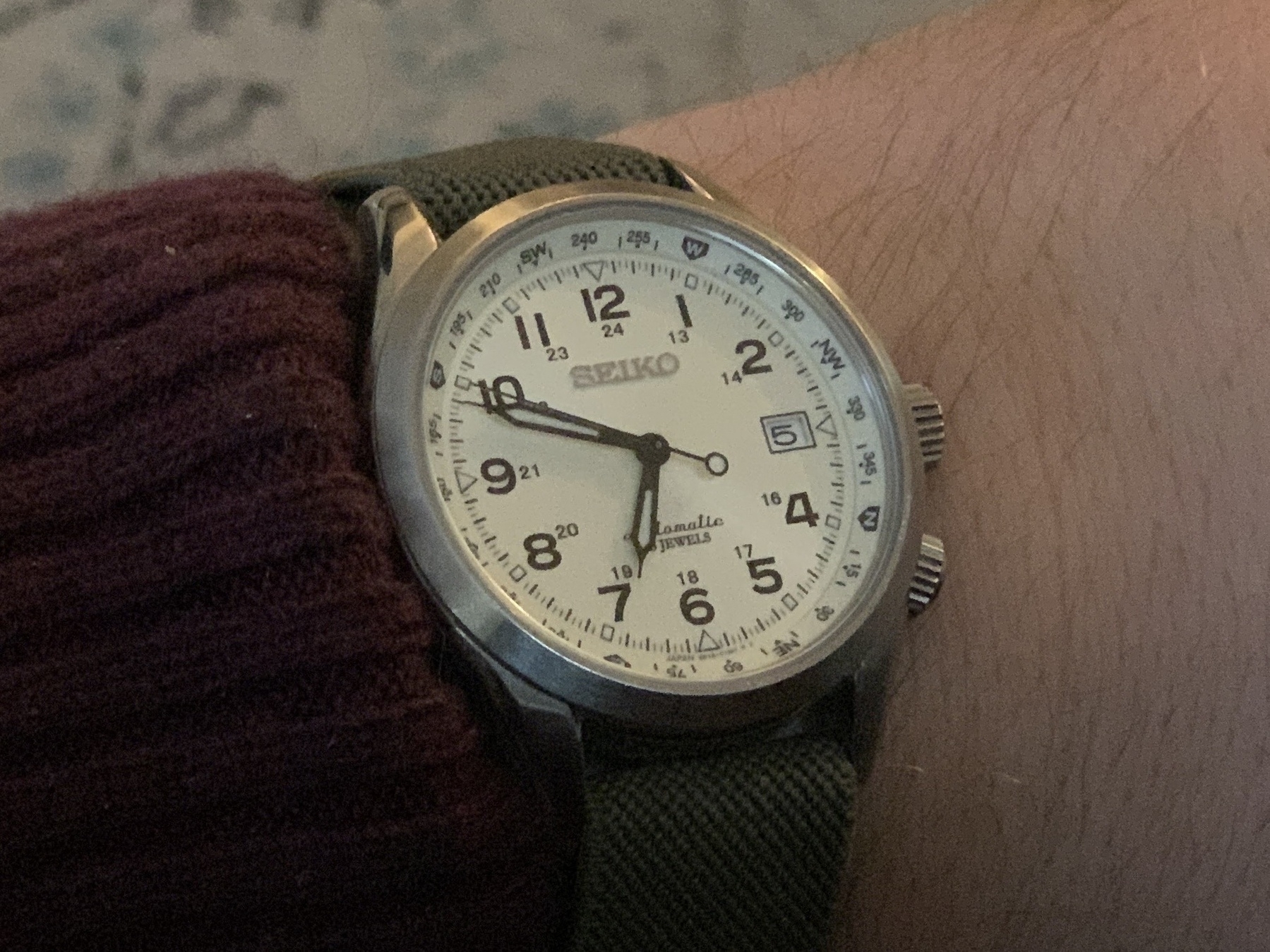 A wristwatch with a green strap, white dial, and black numerals is being worn on a person's wrist.
