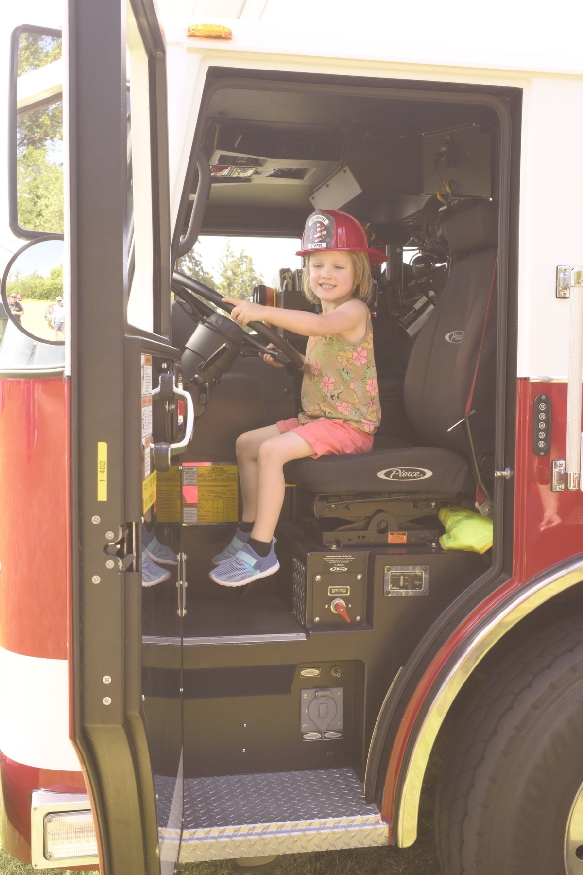 A young child wearing a firefighter helmet is sitting in the driver's seat of a fire truck, smiling.