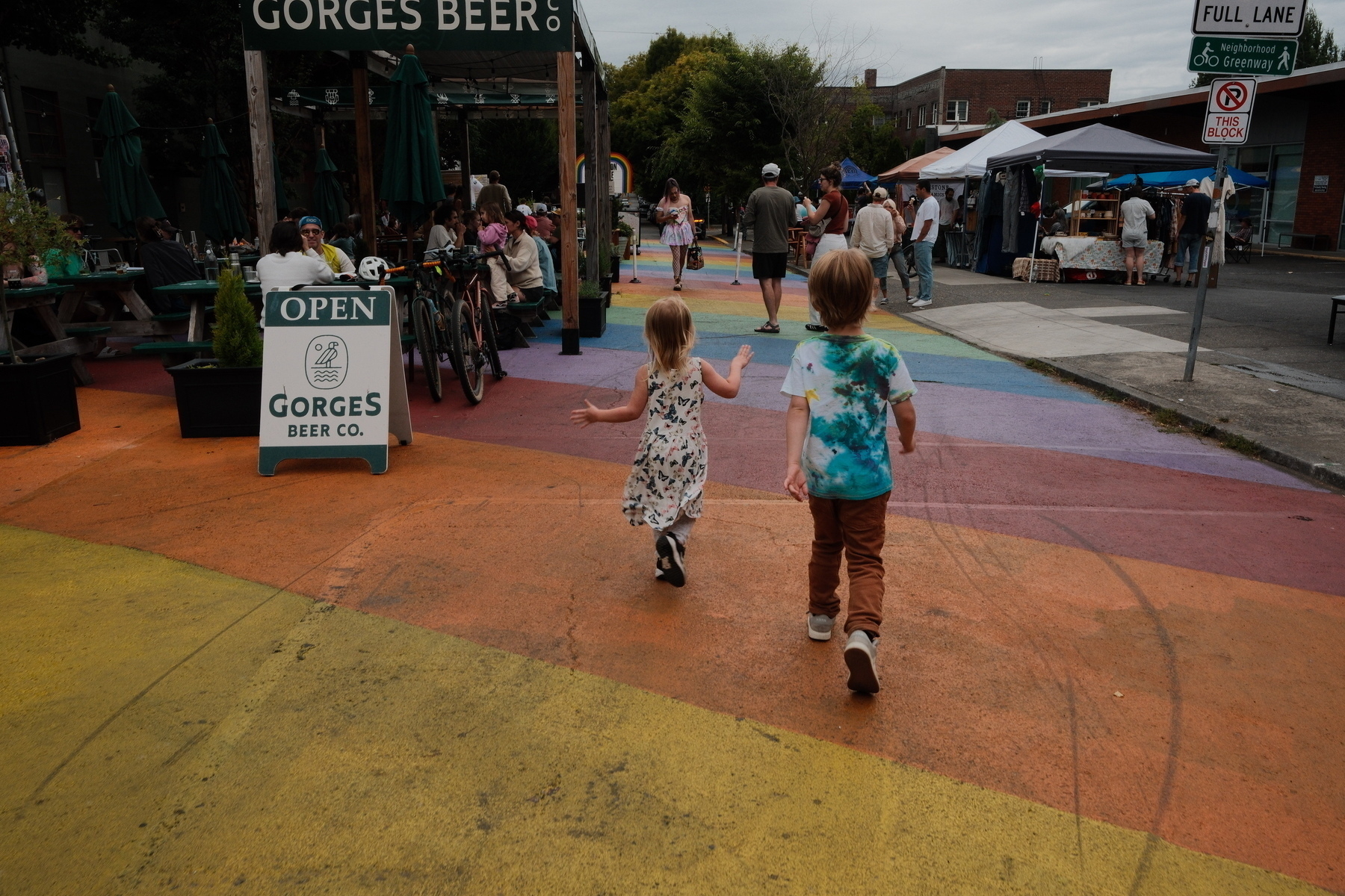 Two children walk on a colorful street toward an outdoor area with tables and people near a sign for Gorges Beer Co.