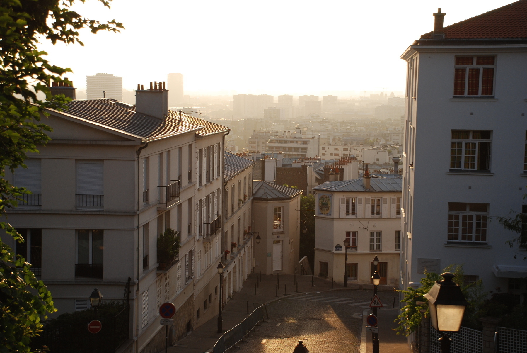 A sunlit street in a city, bordered by residential buildings, descends downhill with trees and distant urban skyline in the background.