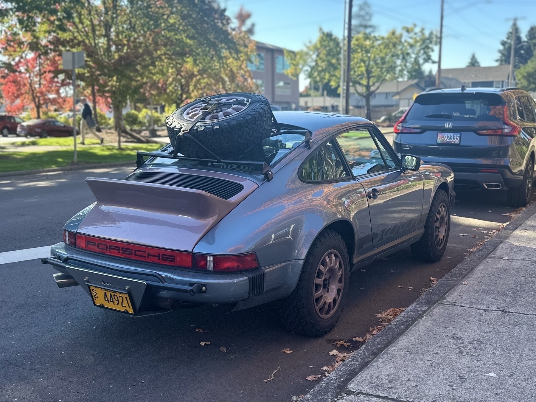 A silver Porsche with a spare tire mounted on its roof is parked on a street next to an SUV and trees with autumn foliage.