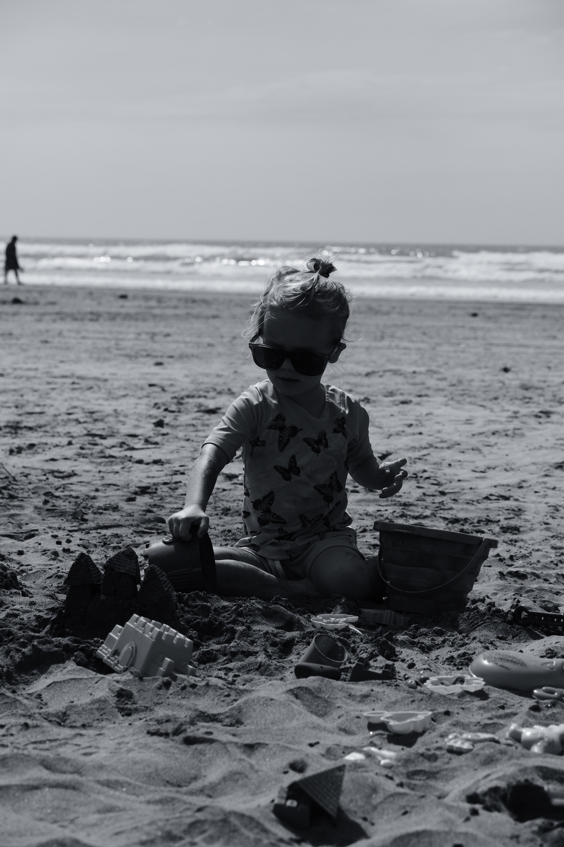 A child is sitting on a beach wearing sunglasses, playing with sand toys, with the ocean and another person in the background.