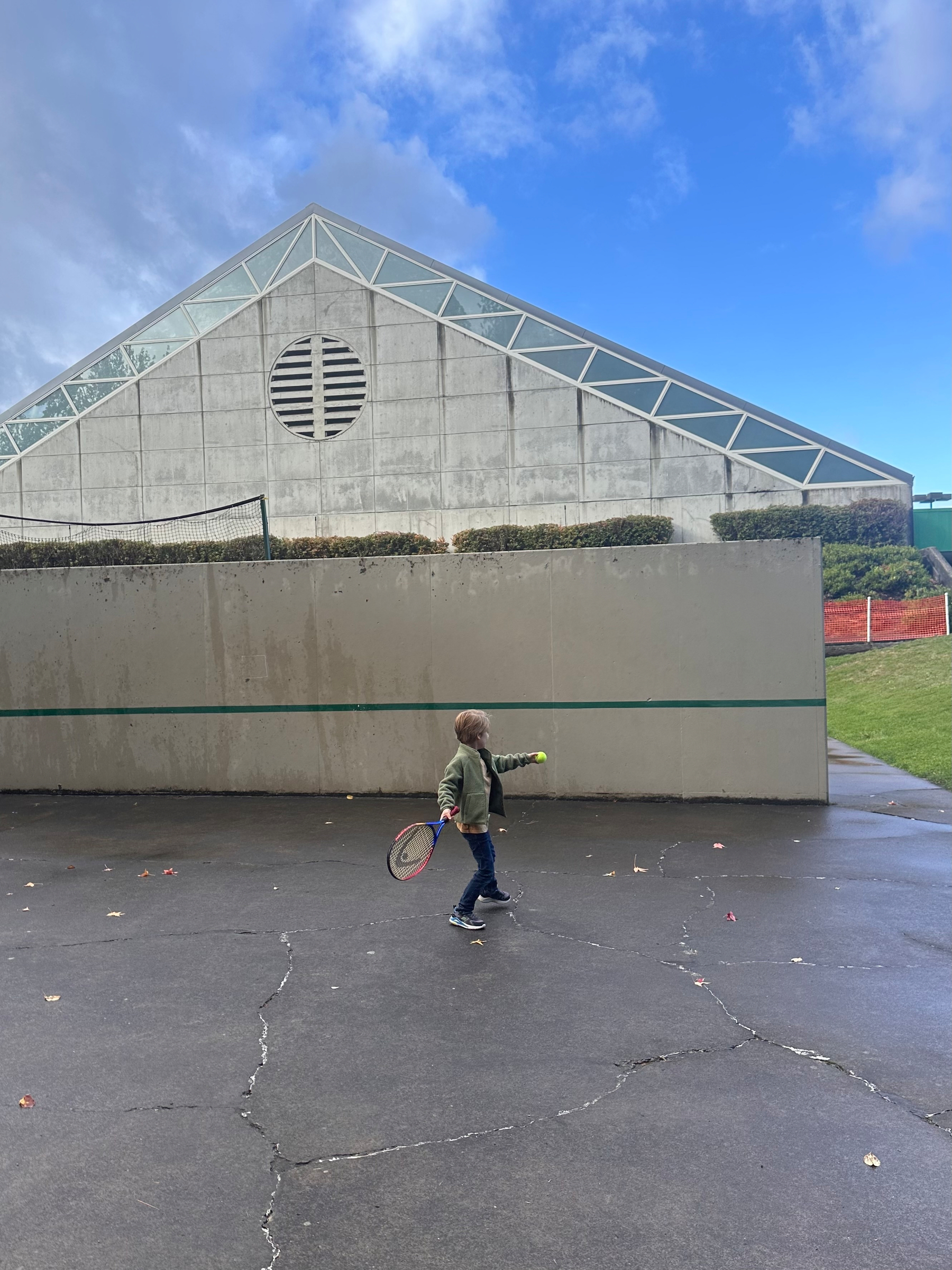 A child holding a tennis racket walks on a paved surface in front of a large, modern triangular building with a cloudy blue sky above.