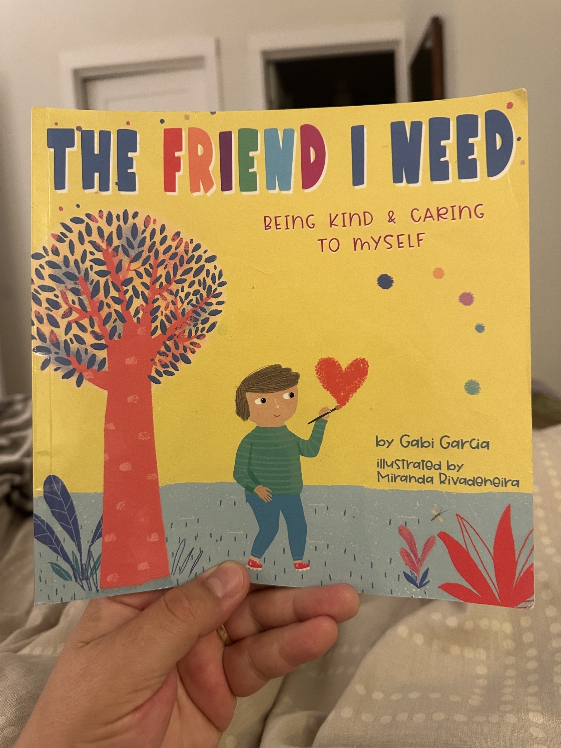 A person holds a children's book titled "The Friend I Need: Being Kind & Caring to Myself" by Gabi Garcia, illustrated by Miranda Rivadeneira, featuring a colorful cover with a drawing of a child and a tree.