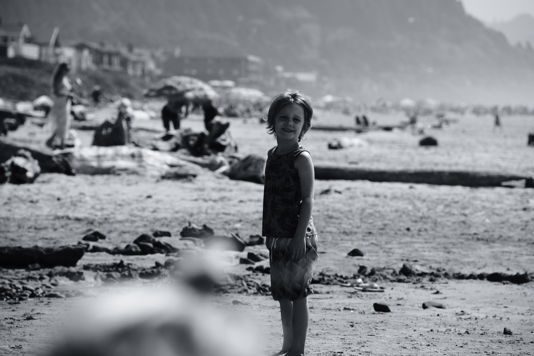 A young child stands on a beach with people and buildings in the background.