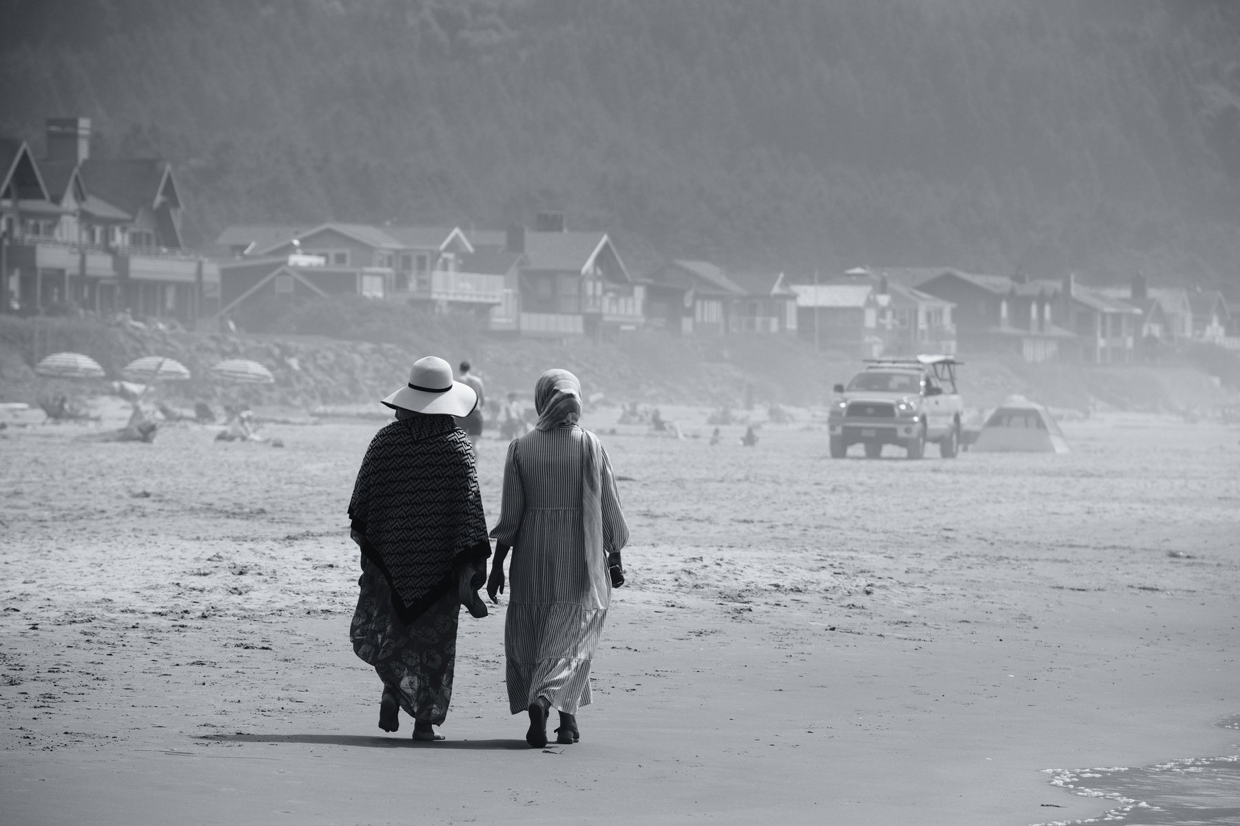 Two people in long dresses and hats walk along a foggy beach with houses and vehicles in the background.