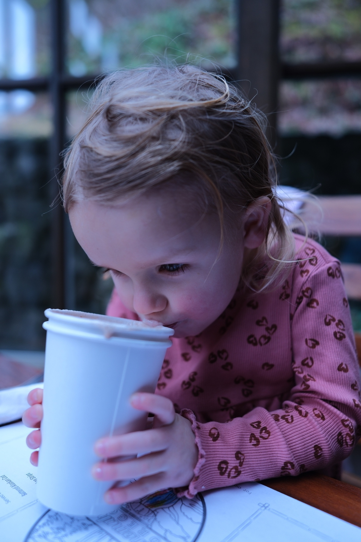 A young child in a pink sweater is drinking from a white cup while sitting at a table.