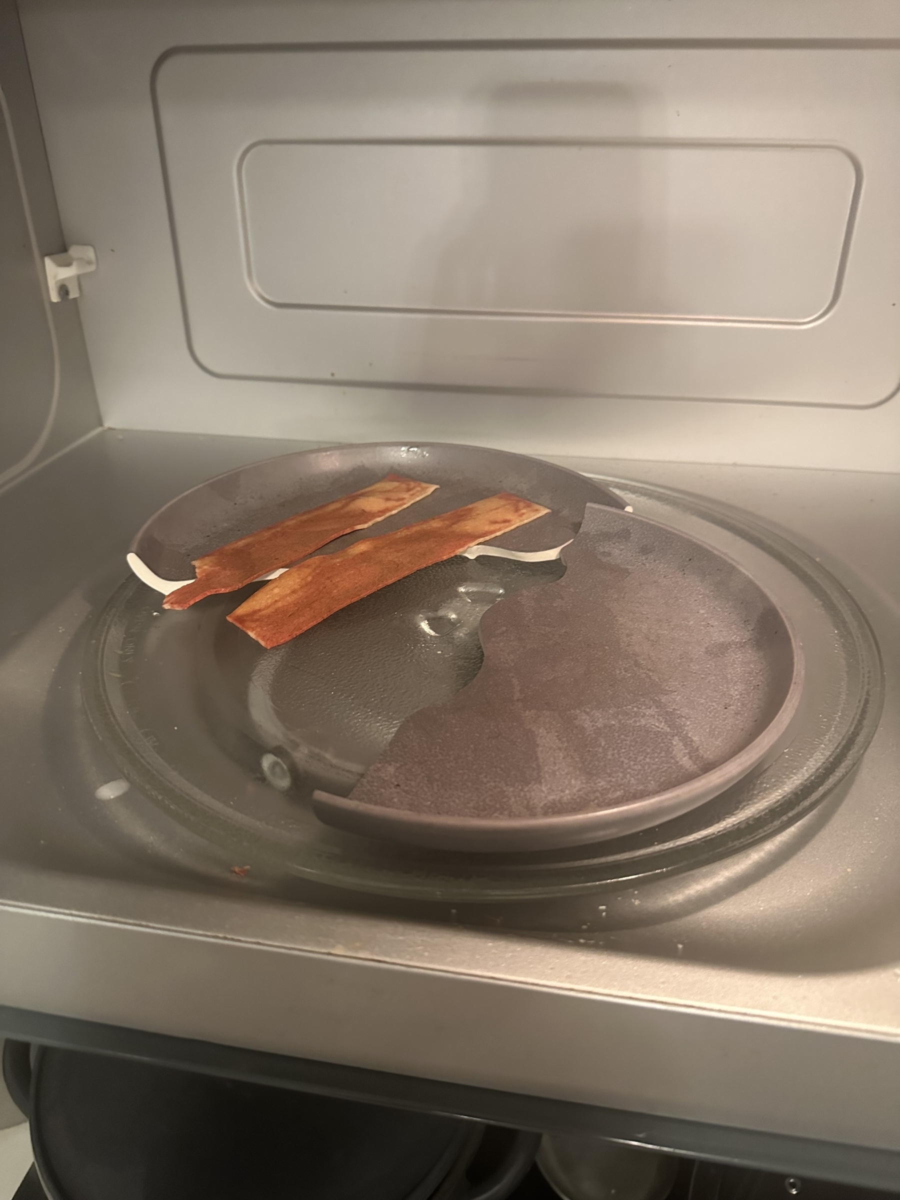 A broken microwave plate with two strips of cooked bacon lies inside a microwave.