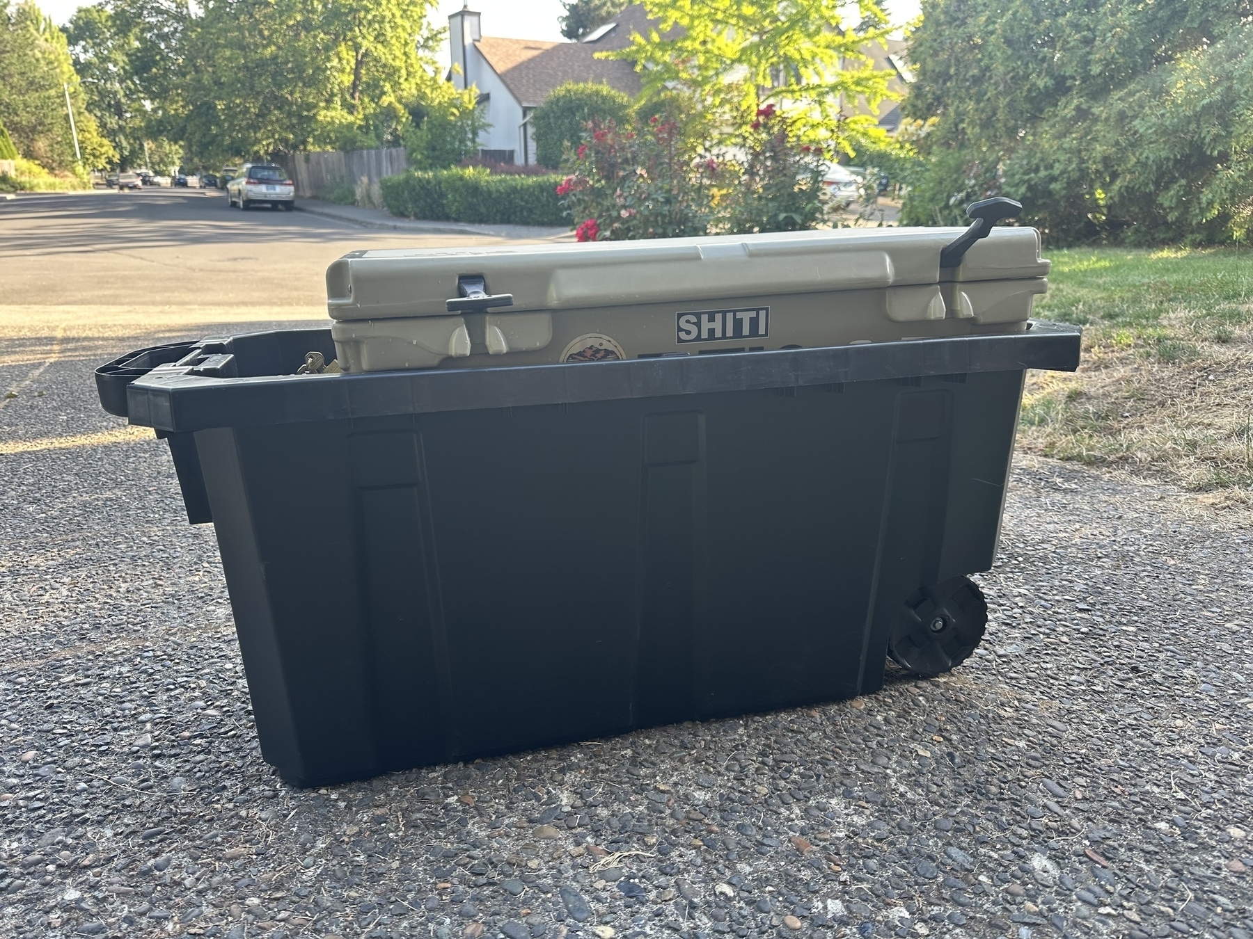 A black plastic storage bin with wheels is placed on a driveway, holding a tan cooler labeled "SHITI."