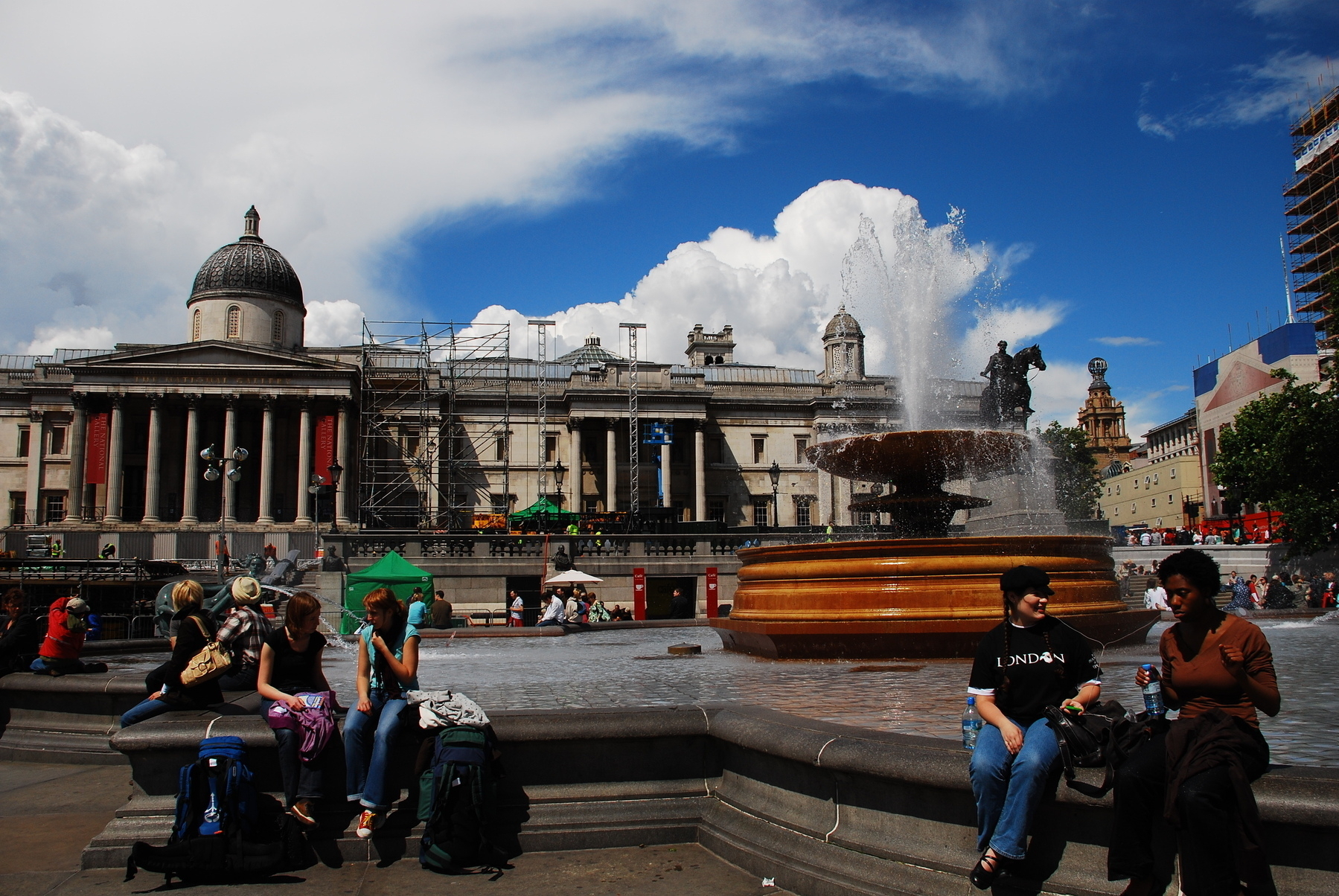 A bustling public square features people relaxing near a large fountain, with a historic building in the background under a blue sky with clouds.