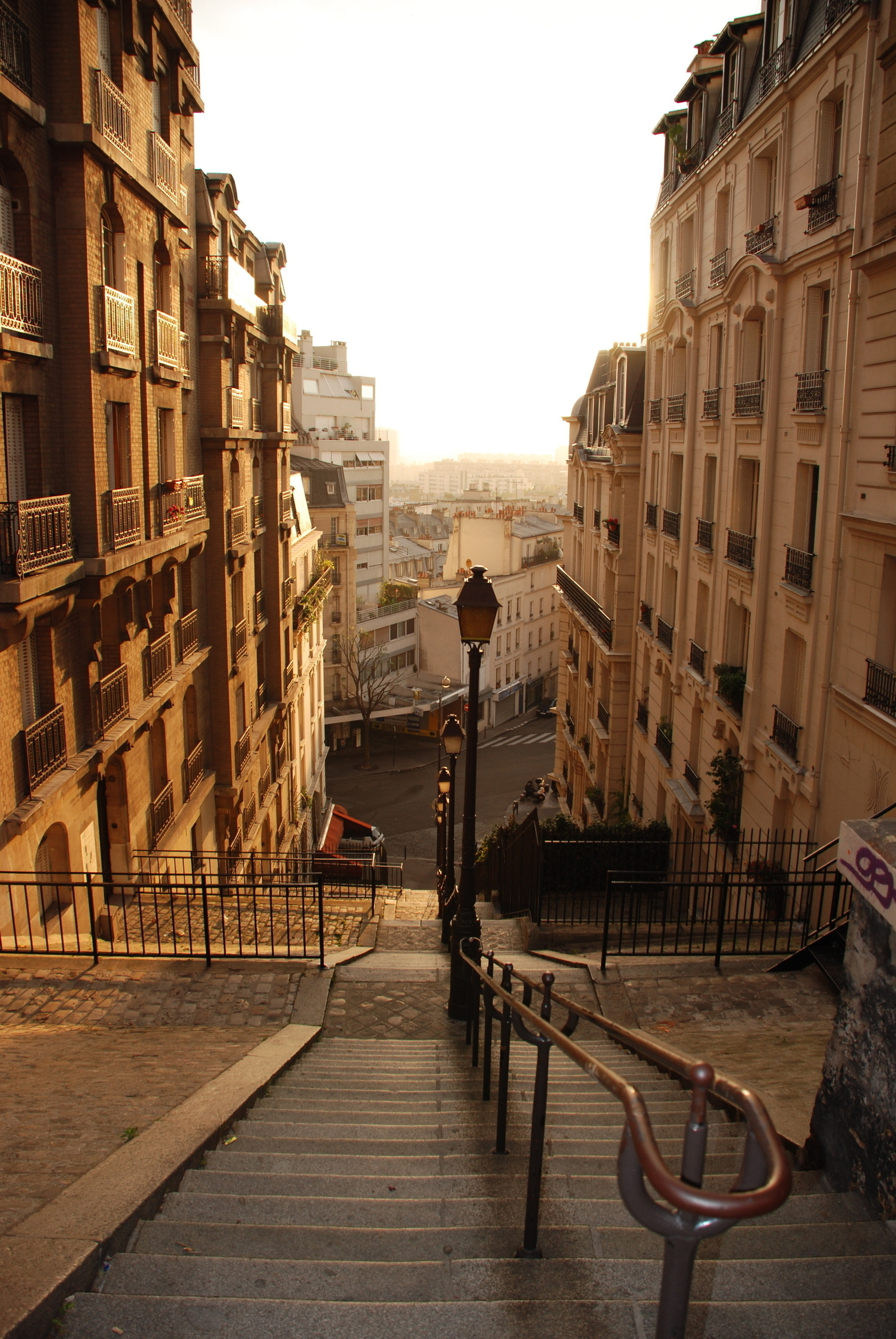 A sunlit, narrow European street flanked by tall, historic buildings features a descending staircase leading to an intersection.