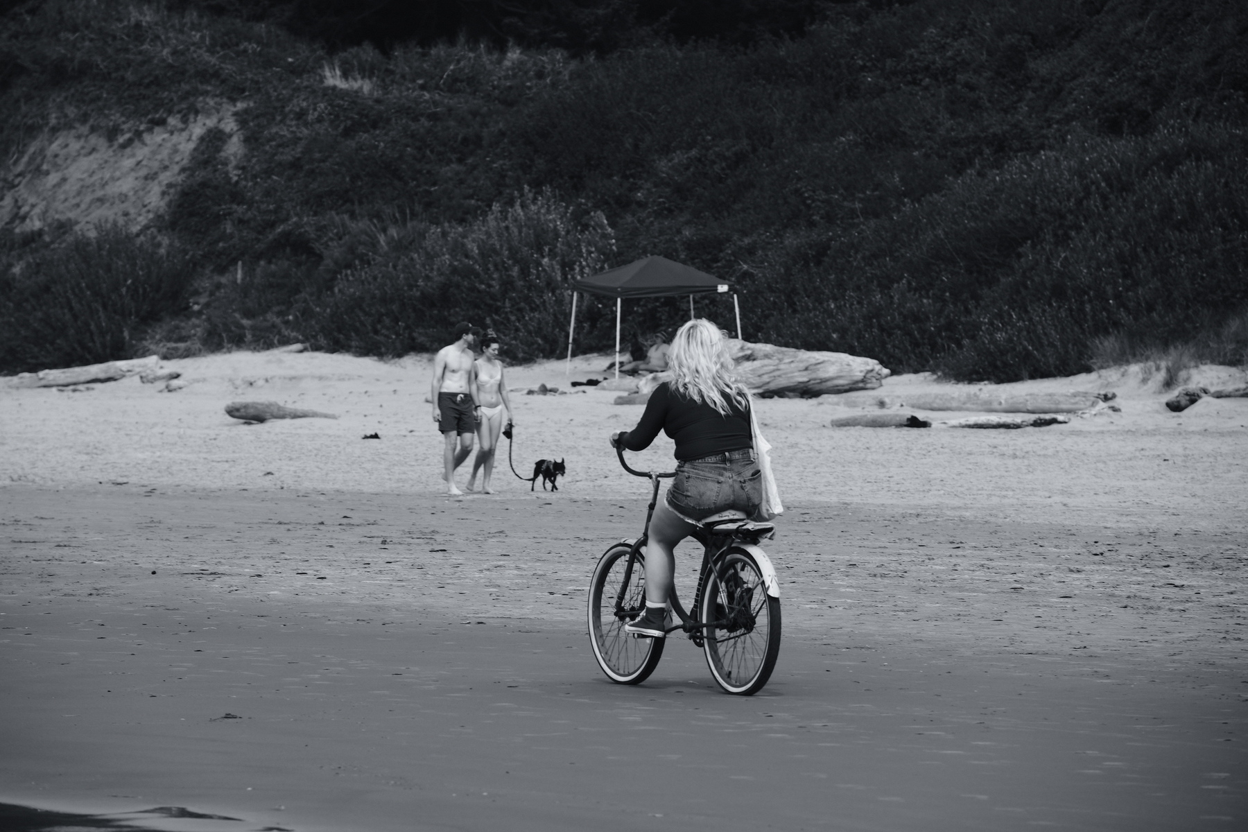 A person is riding a bicycle on a beach while another person walks with a dog near a tent.