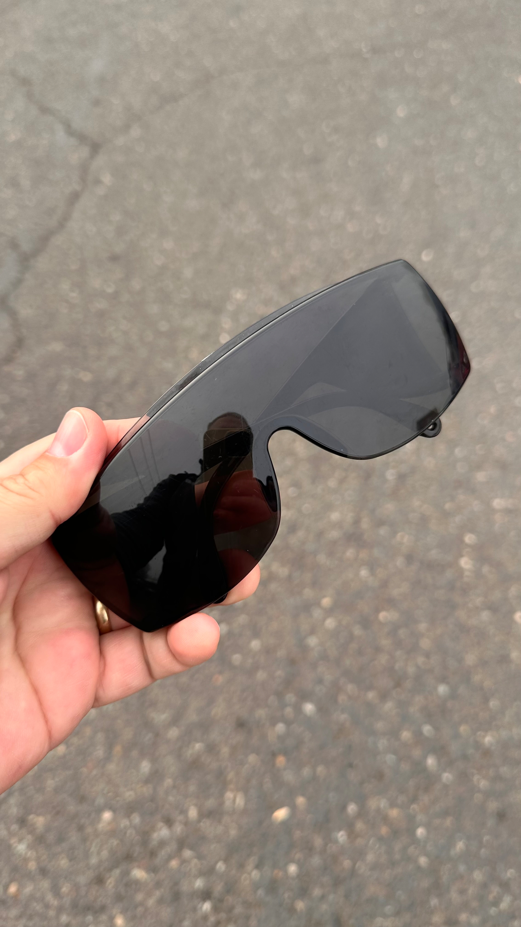 A hand is holding a pair of black, wraparound sunglasses against a background of asphalt.