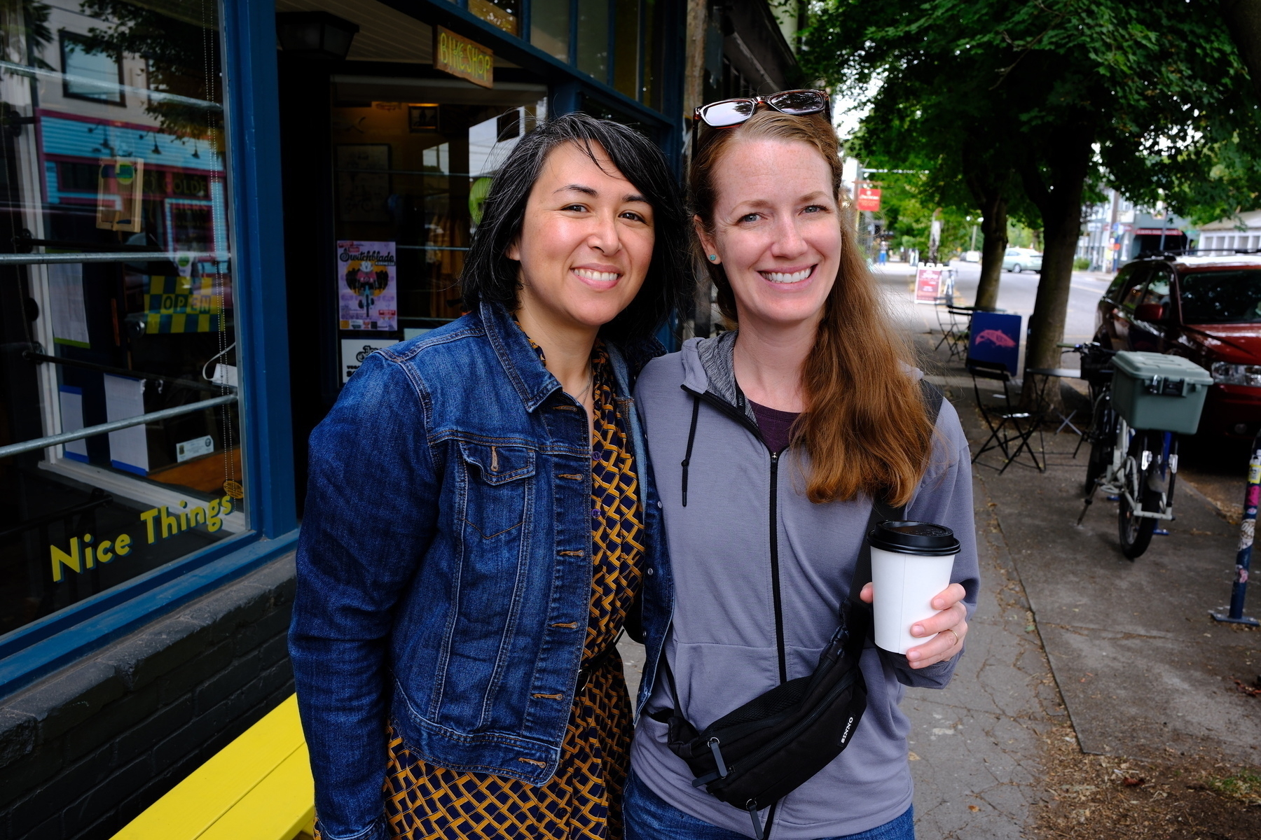 Two people are smiling and standing on a sidewalk, one holding a cup of coffee.