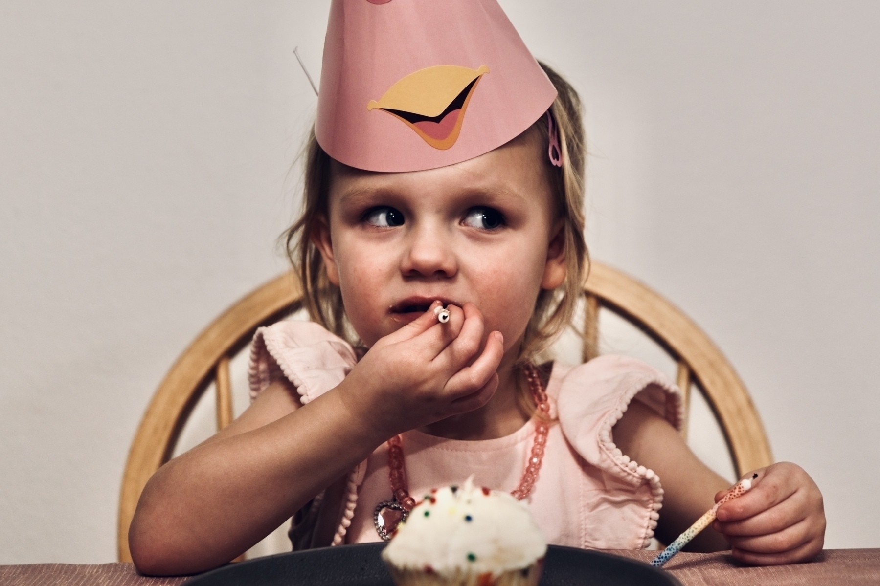 A young child wearing a pink party hat is sitting at a table with a cupcake in front of them.