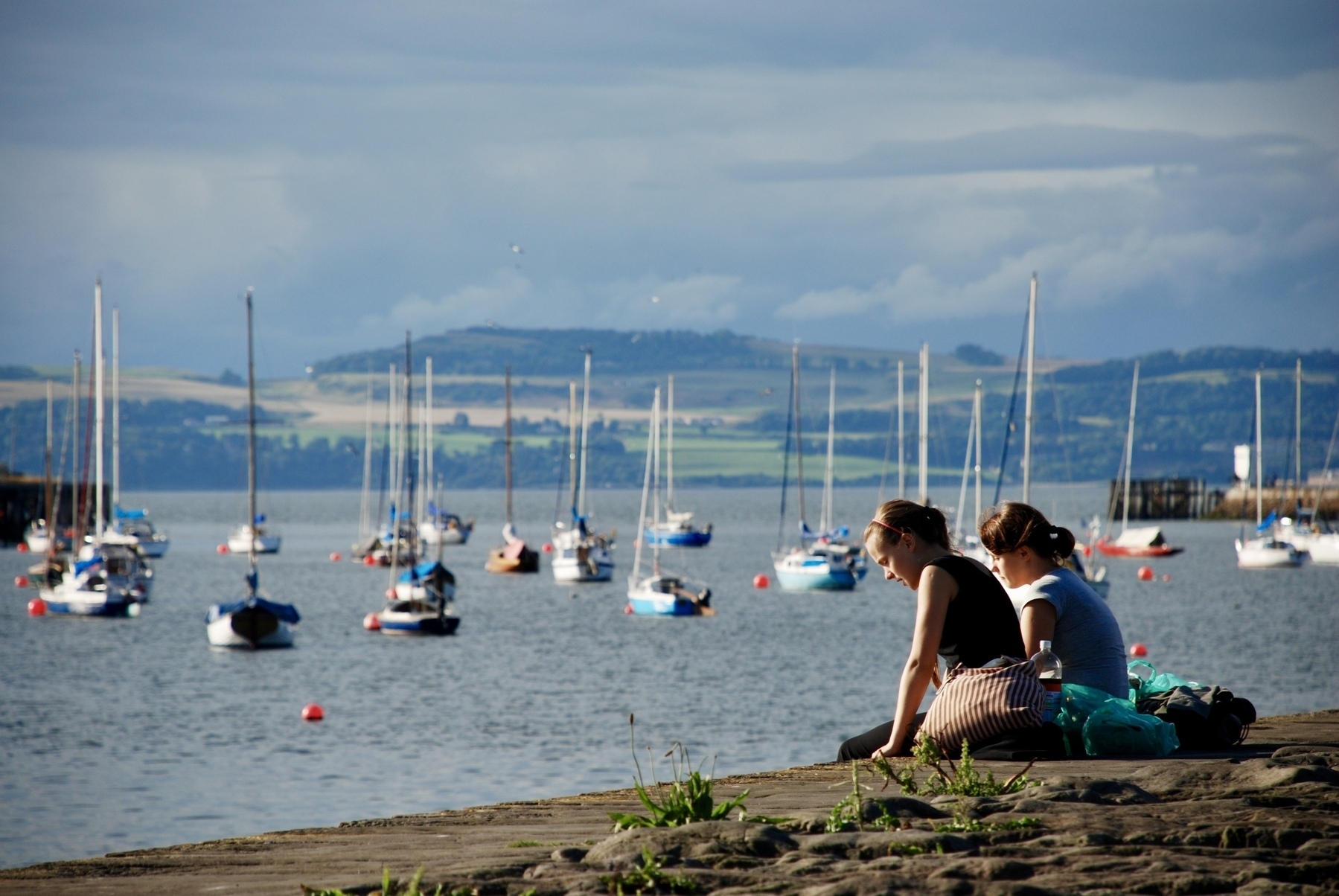 Two people sit on a stone ledge overlooking a calm bay with numerous sailboats anchored in the water. Firth of Forth, Scotland. 