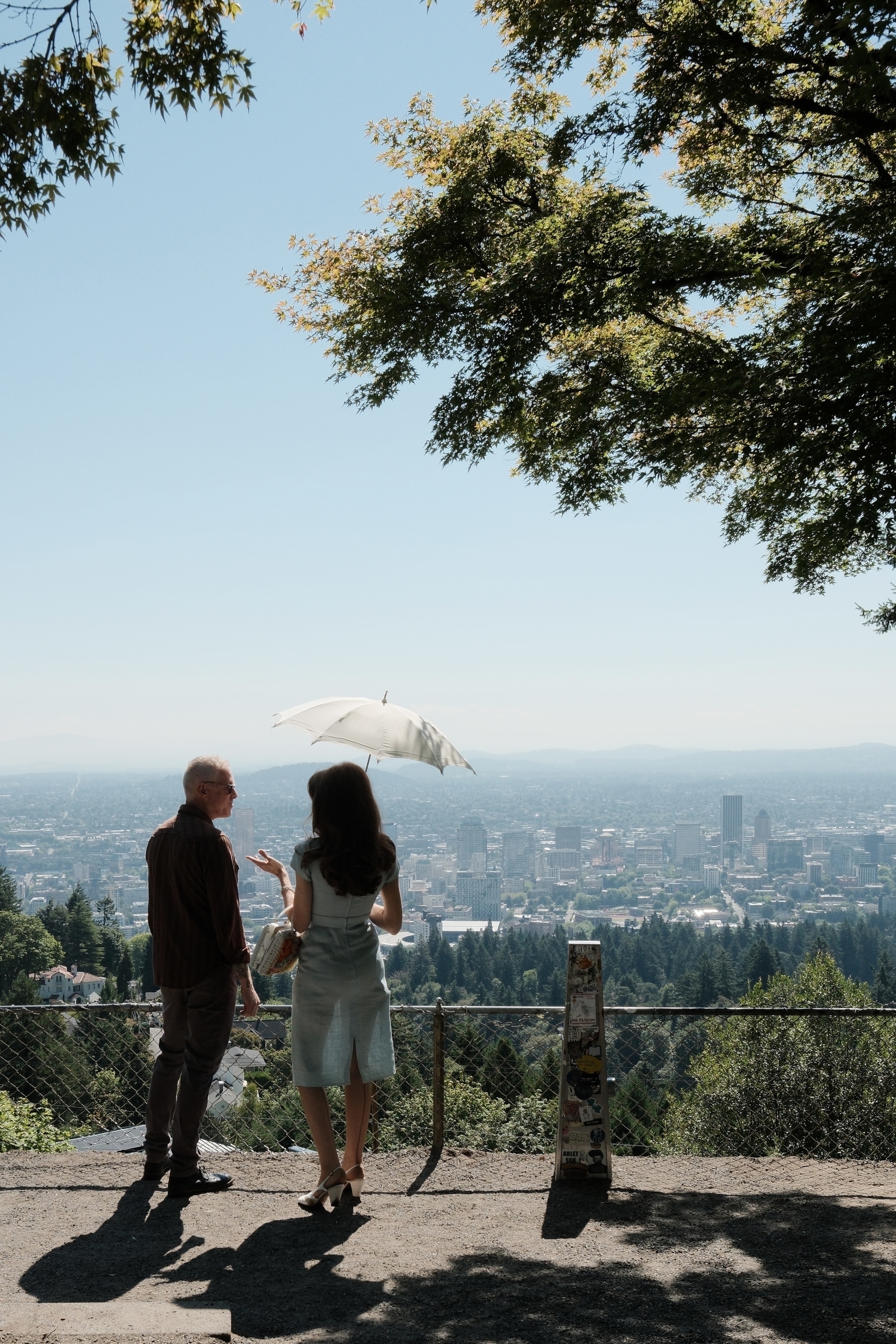 Two people stand on a scenic overlook with one holding an umbrella, gazing at a cityscape surrounded by greenery under a clear sky.