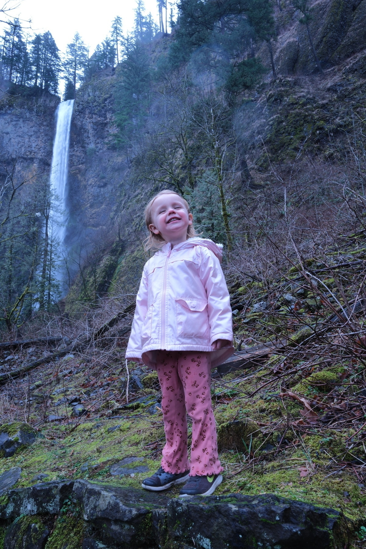 A smiling child in a pink outfit stands on a mossy surface with a waterfall and forested hills in the background.