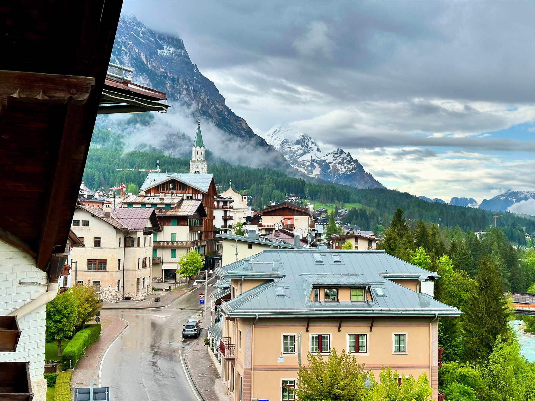 A picturesque alpine village is set against a backdrop of towering snow-capped mountains under a cloudy sky.