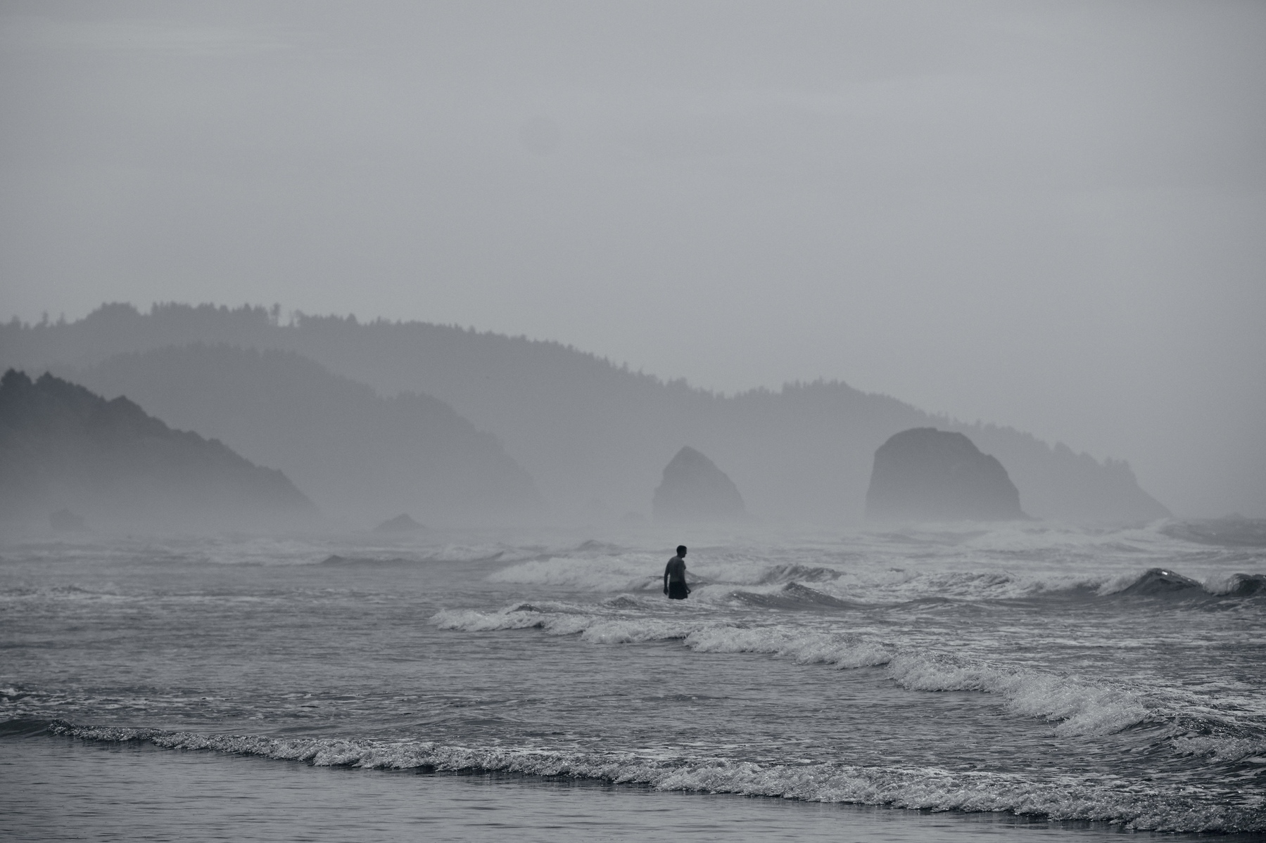 A lone figure stands in the water, surrounded by misty waves and distant rocky outcrops under a gray sky.
