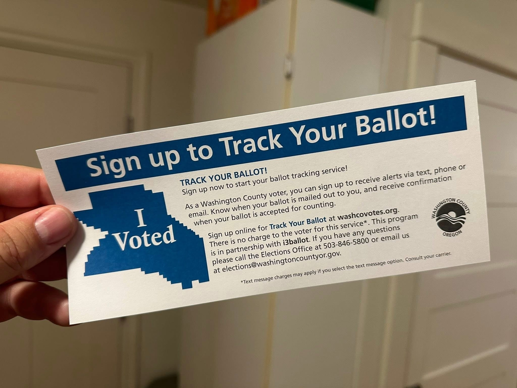 A hand is holding a flyer instructing how to sign up for a ballot tracking service for Washington County voters, featuring a blue I Voted sticker graphic.