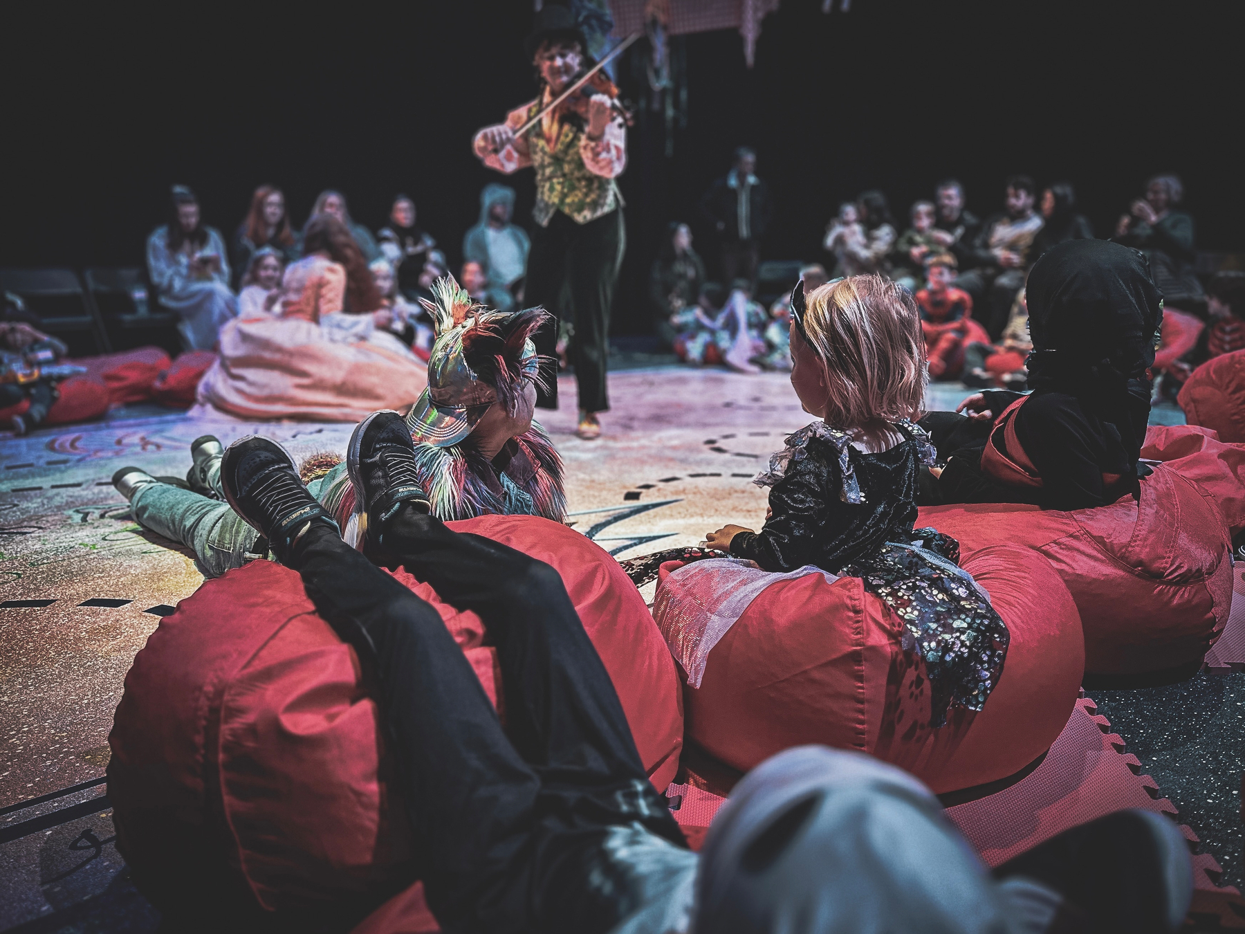 Auto-generated description: A performer plays the violin to an audience of children in costumes sitting on red bean bags.