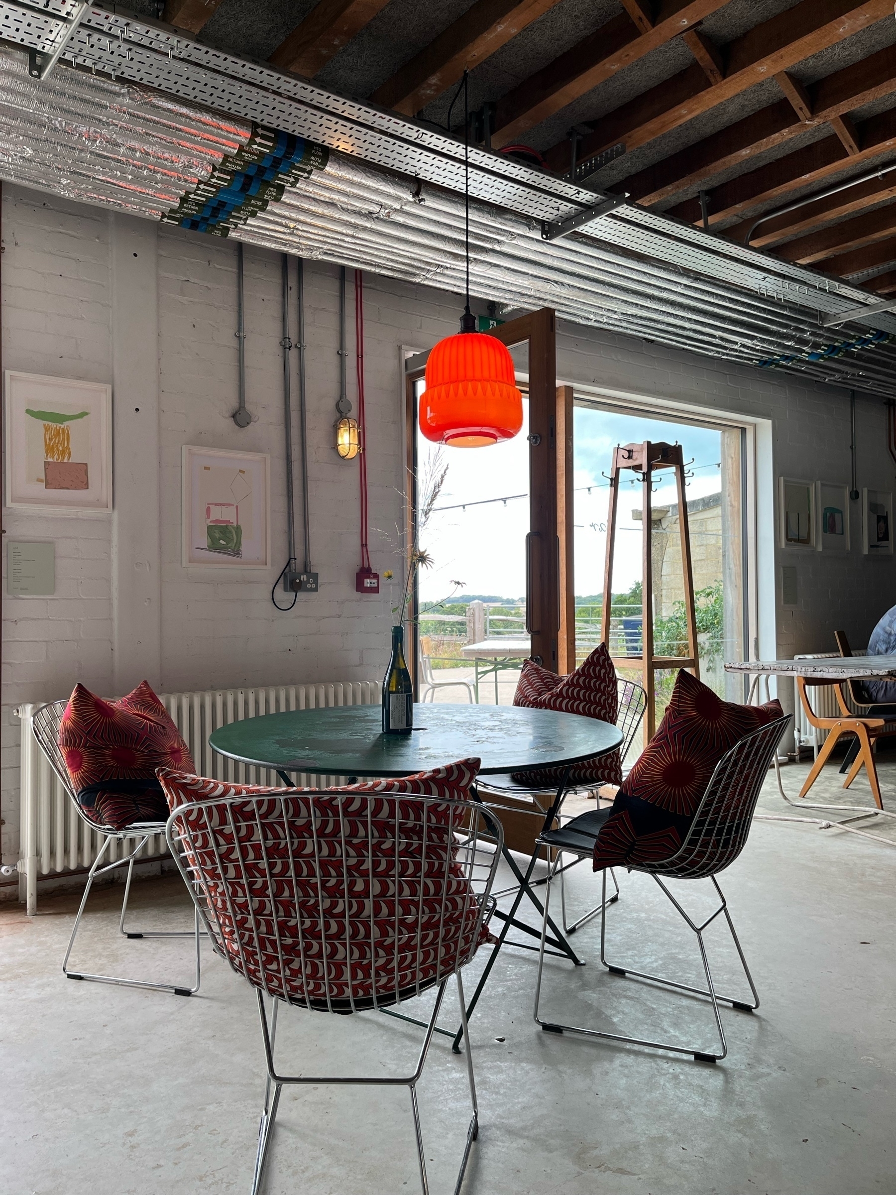 Green round table with orange lamp hanging from the ceiling, four chairs around it with red cuisons on an industrial setting